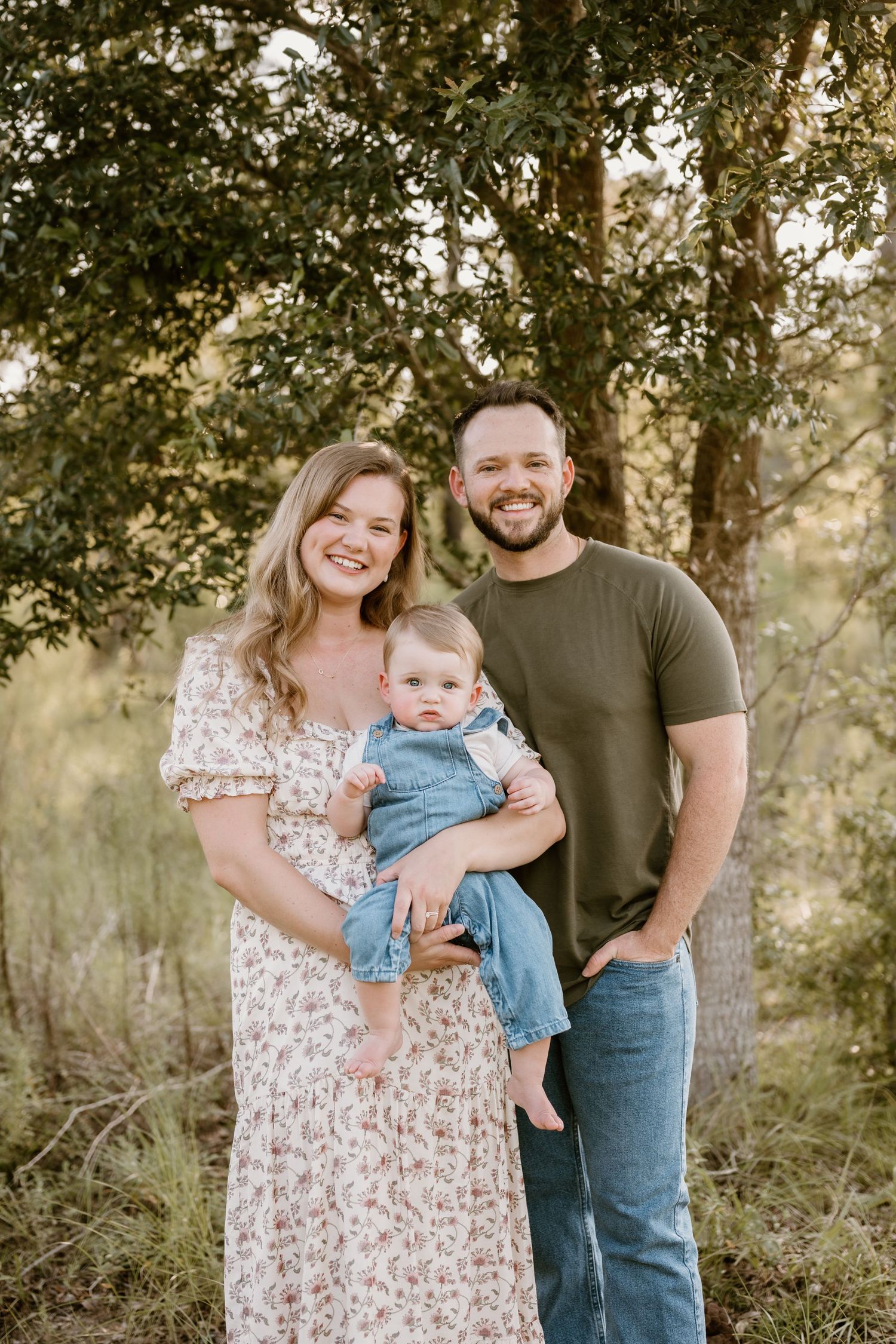 Family portrait outdoors with baby in denim overalls against tree backdrop.