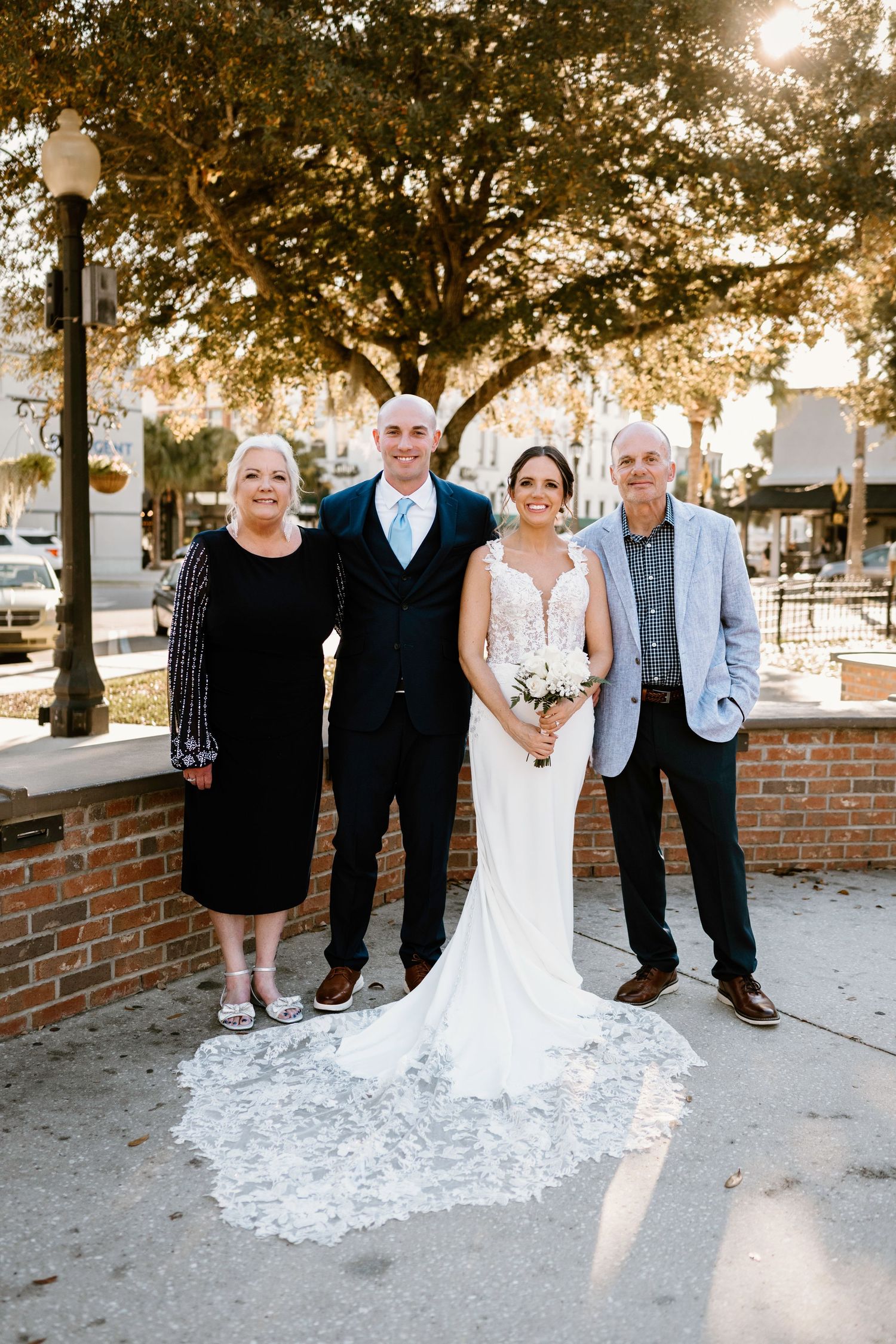 Wedding party poses together outdoors with brick wall and trees in golden sunlight.
