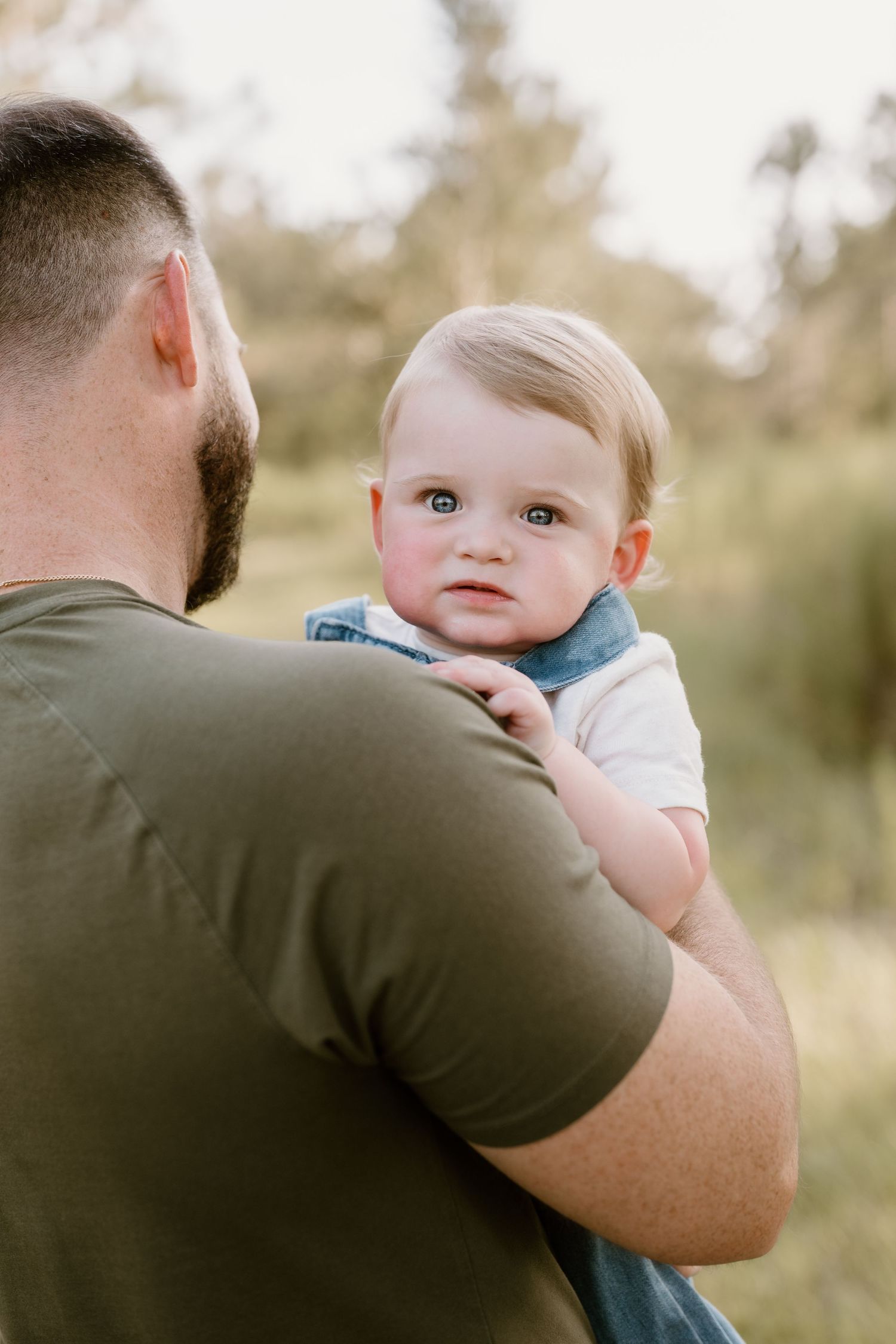Baby being held by parent in olive green shirt against blurred natural background.