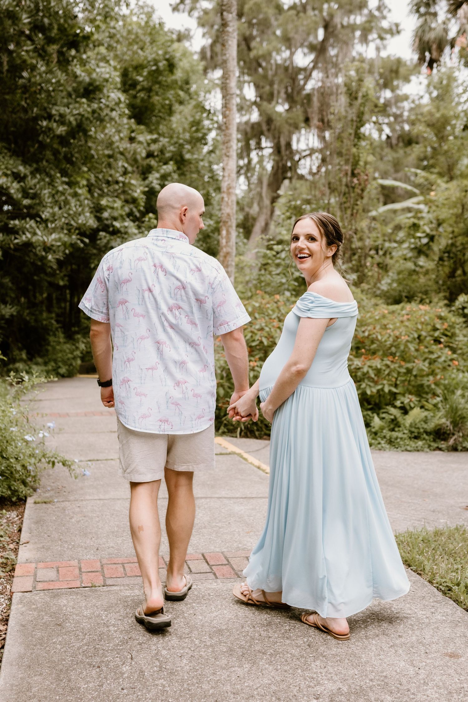 A couple in coordinating outfits walk together on a garden path surrounded by lush greenery.