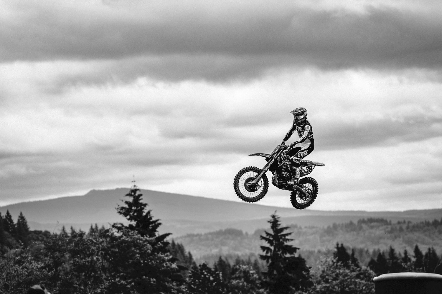 Black and white photo of a motocross rider performing a high jump on a dirt bike against a cloudy sky.