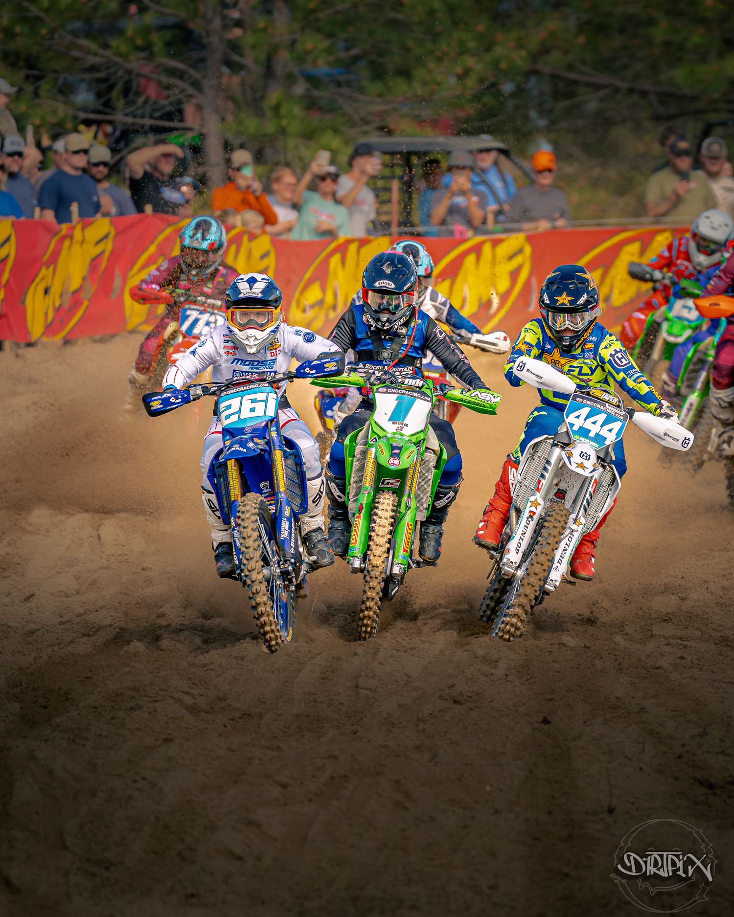 Group of motocross riders racing side by side at start of dirt track competition.
