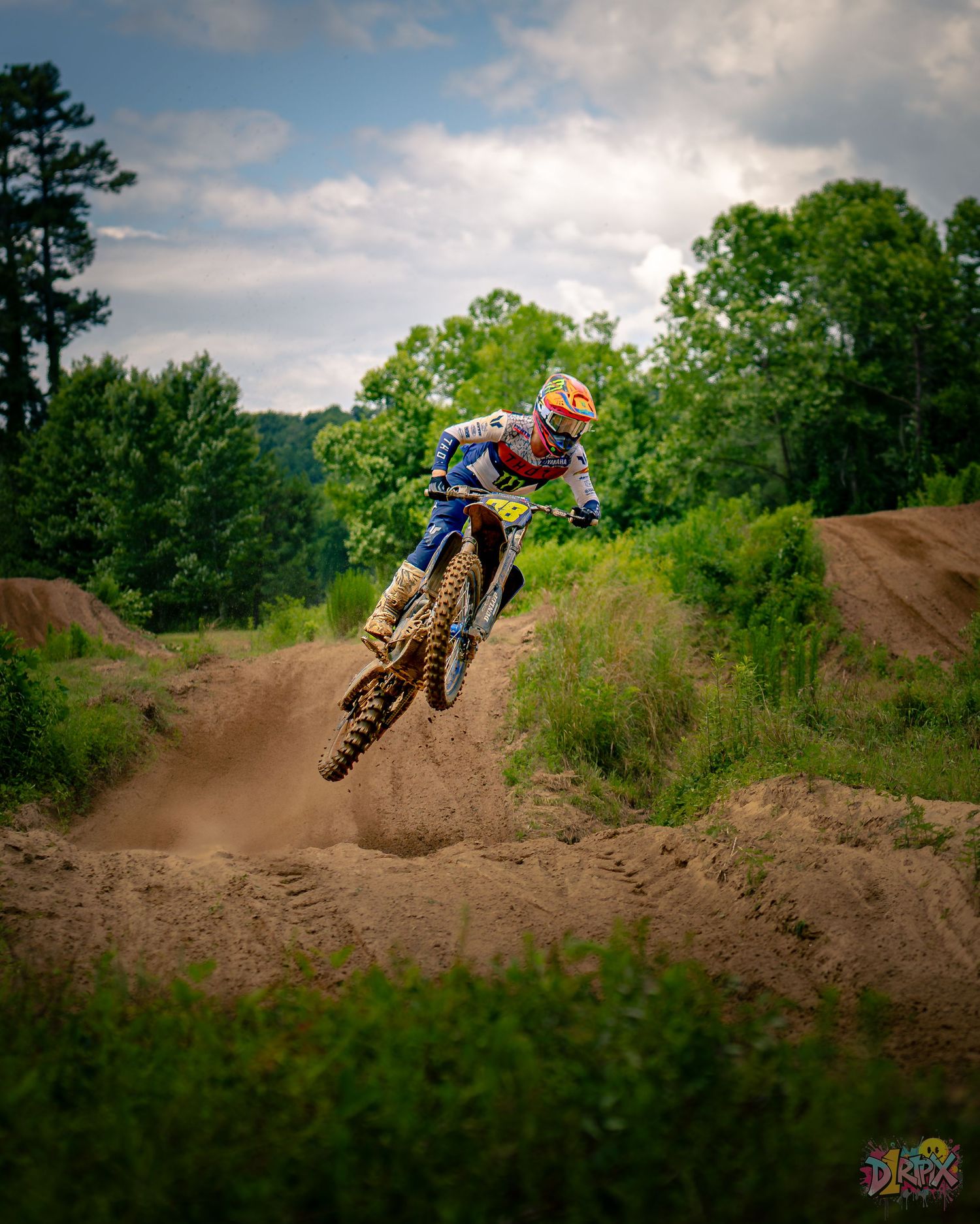 Dirt bike riders racing over multiple jumps on outdoor motocross track with trees in background.