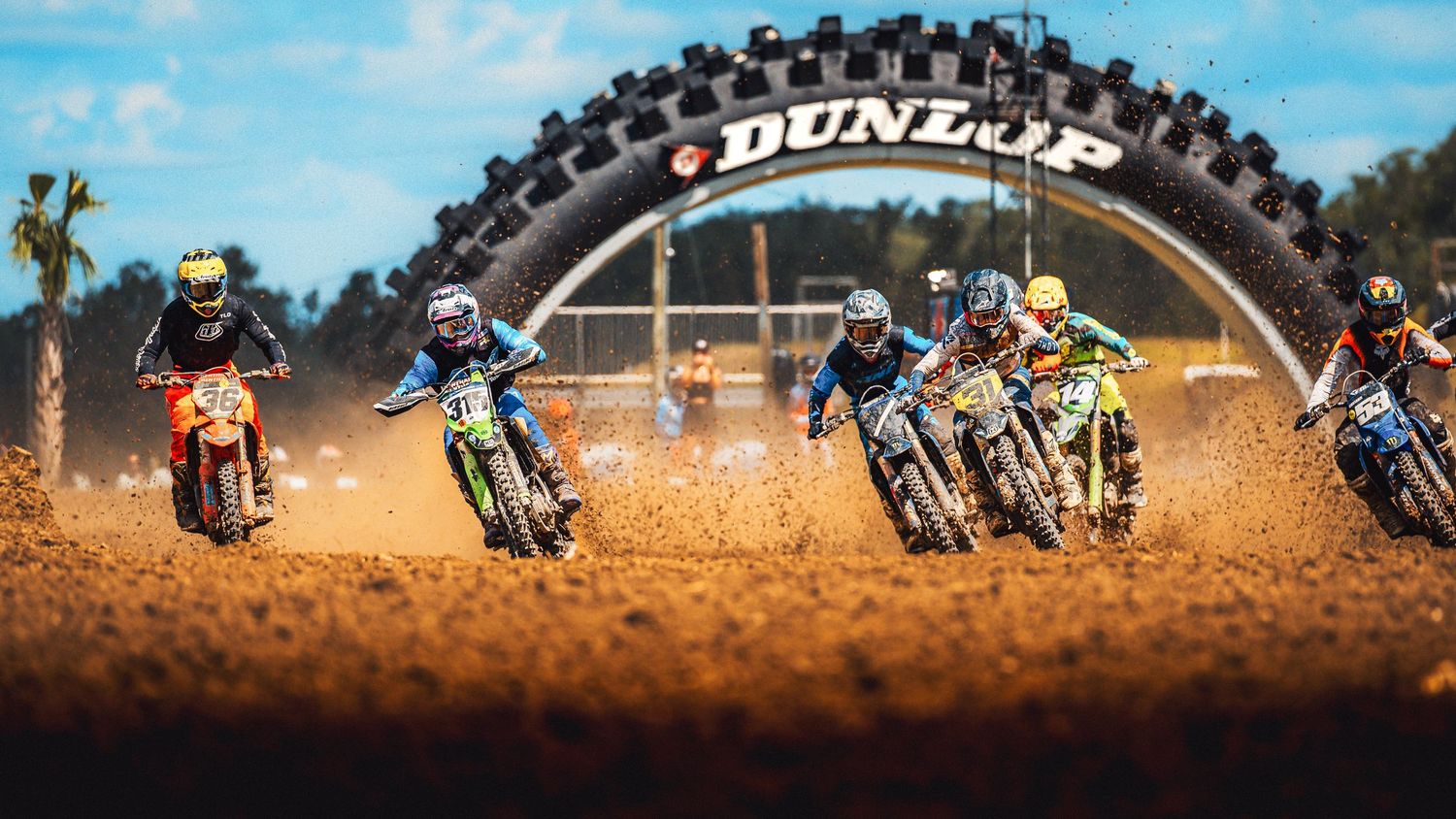 Motocross riders race on a dirt track under a Dunlop tire arch at a professional off-road motorcycle competition.