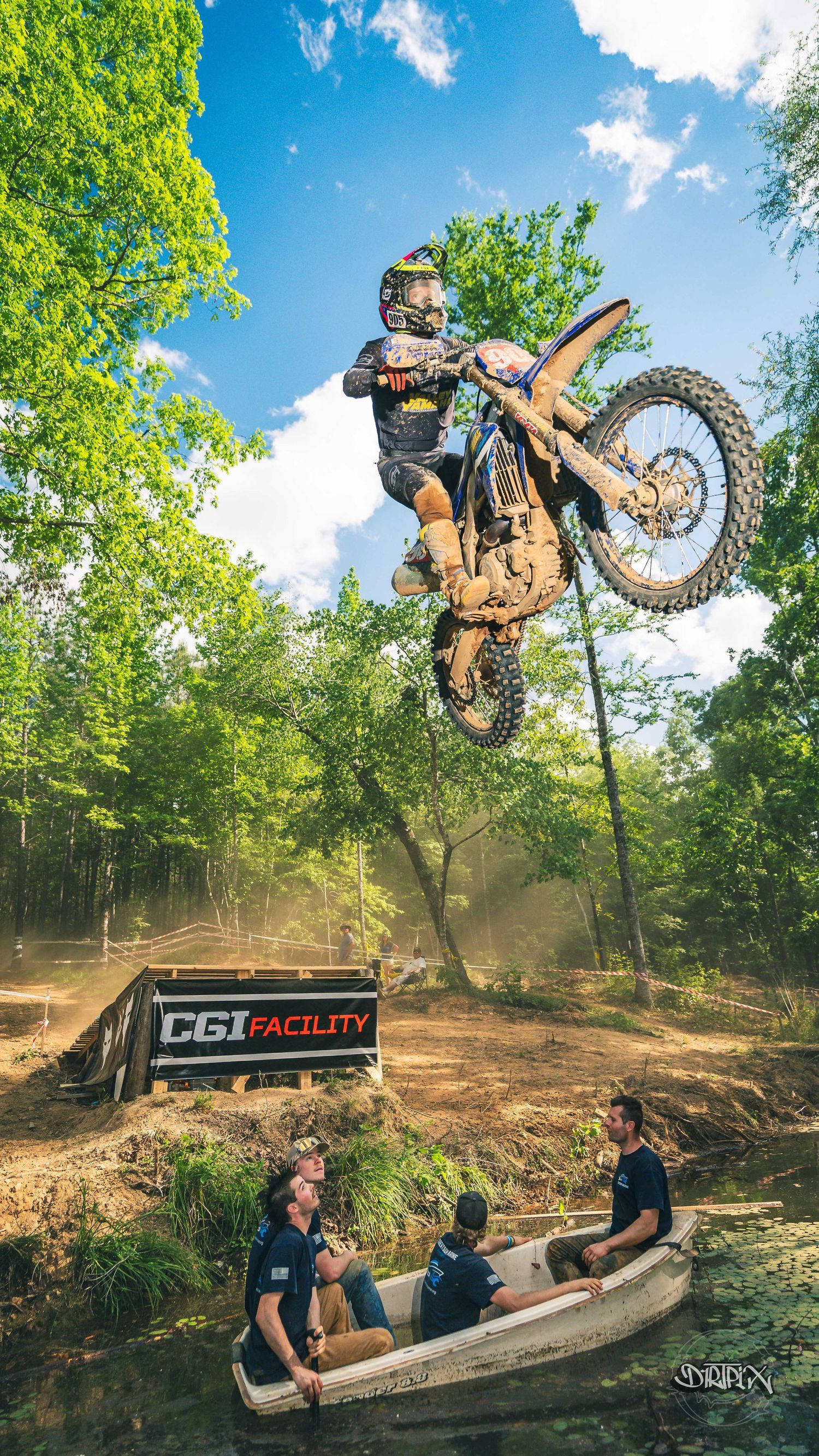 Motocross rider performs a high jump over a boat with spectators at CGI Facility outdoor track surrounded by trees.