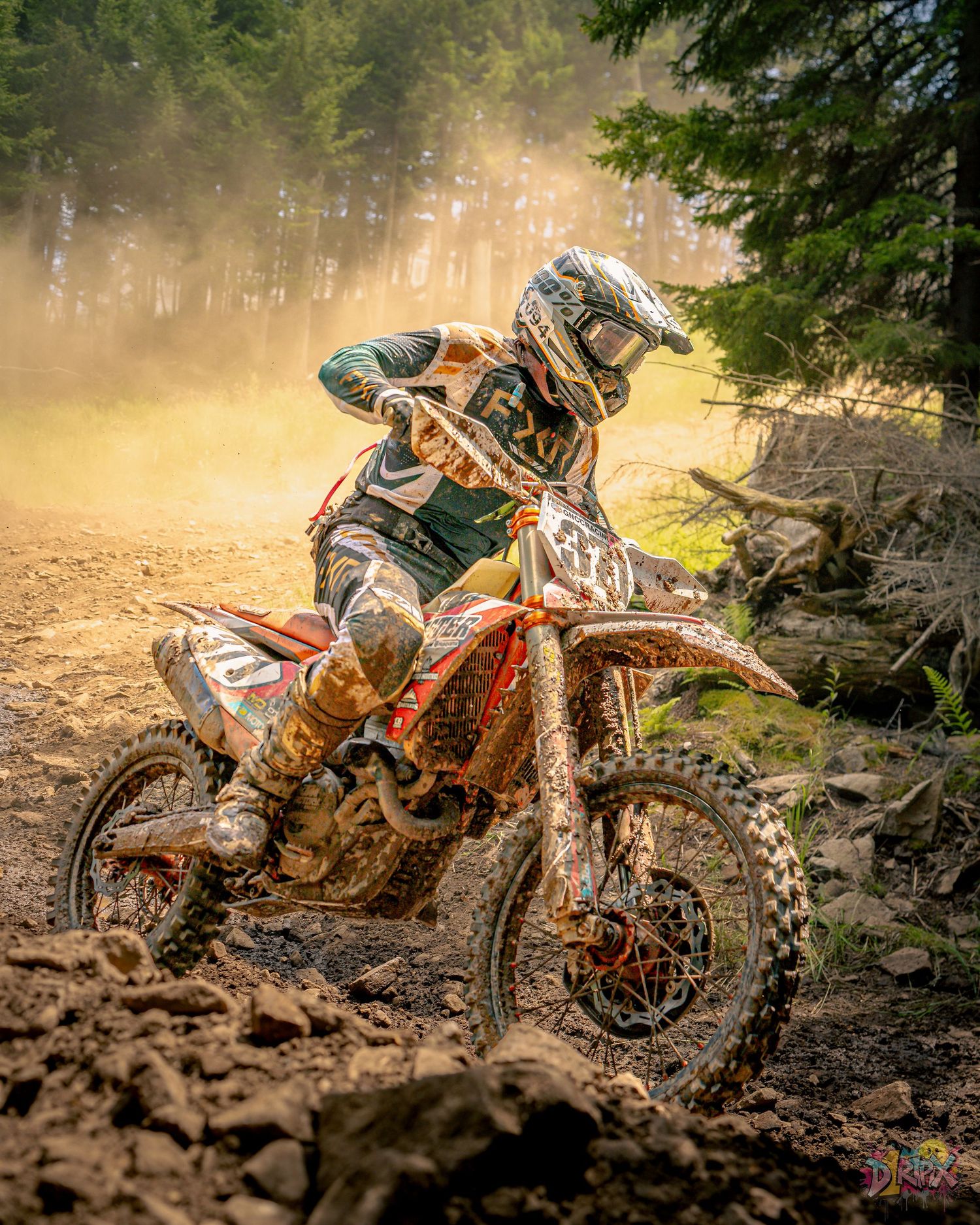 Dirt bike rider navigates through muddy forest terrain in dramatic lighting conditions during off-road competition.