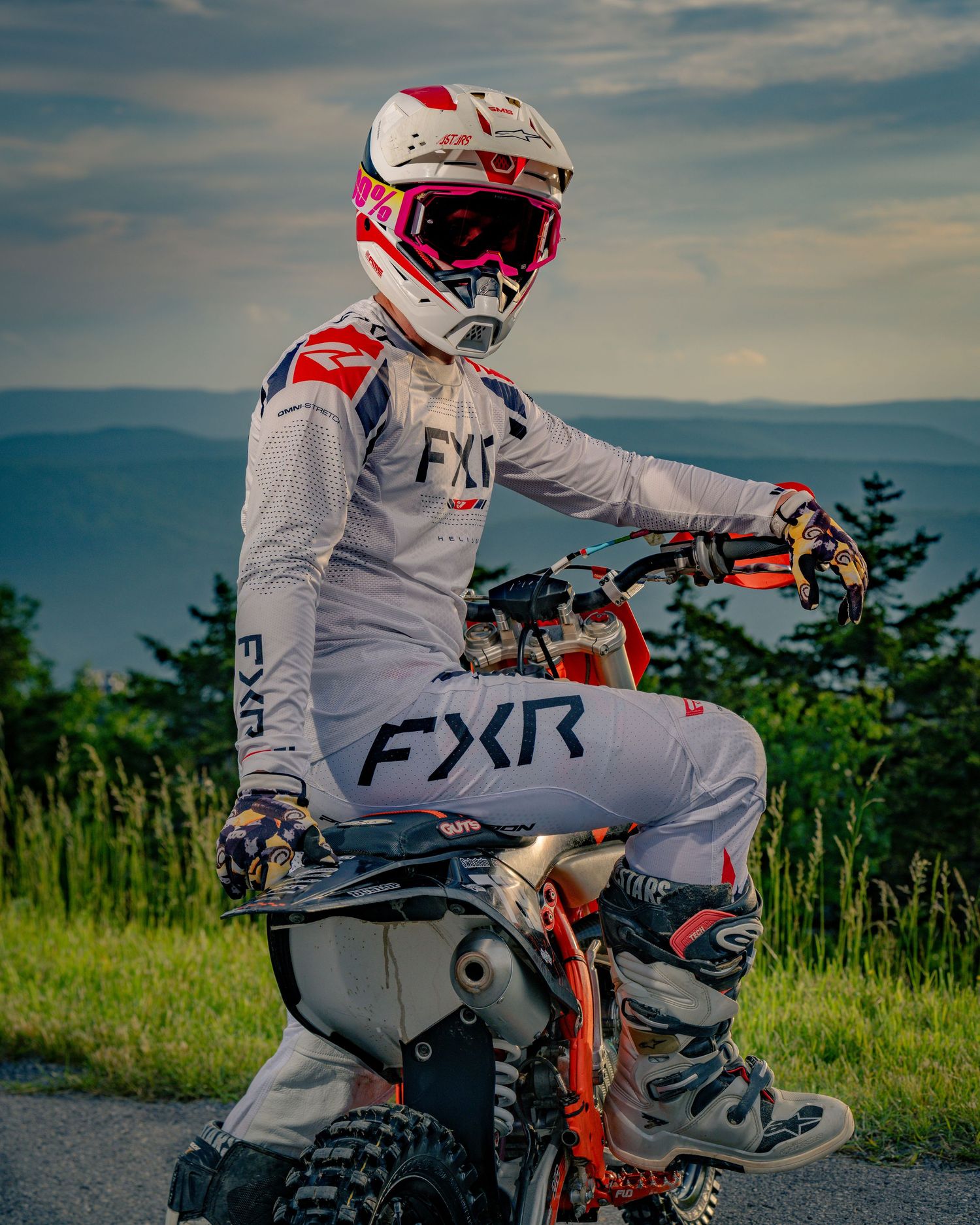 Dirt bike rider in gray FXR gear sitting on motorcycle against scenic landscape.