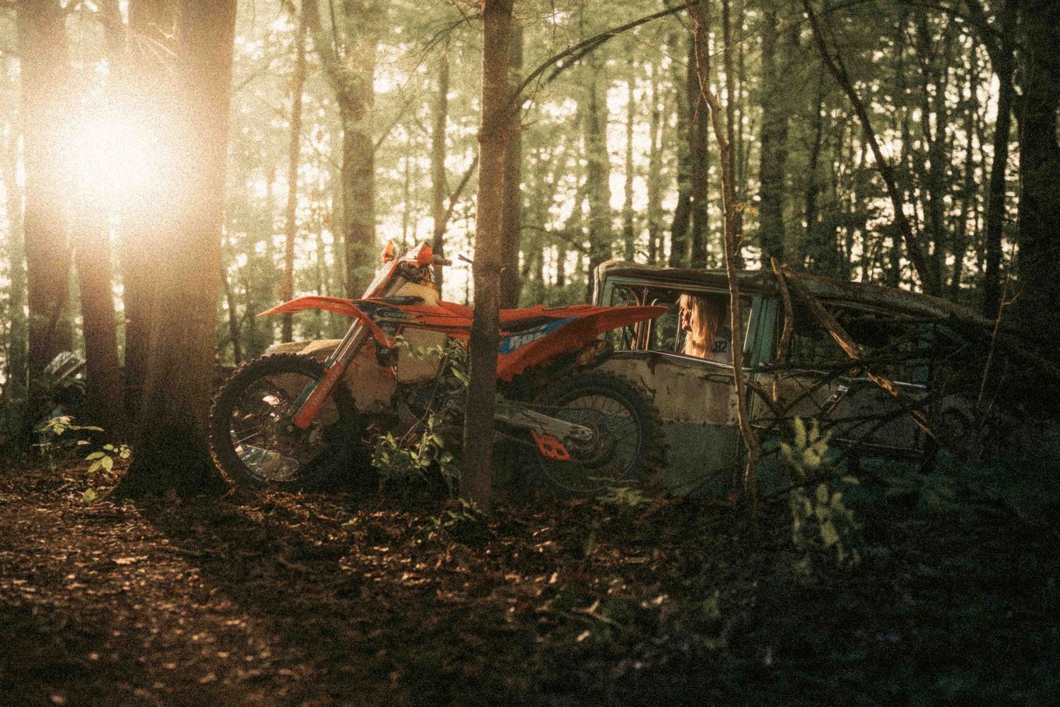 Orange dirt bike parked against a rusted abandoned truck in a sunlit forest with rays of light filtering through trees.