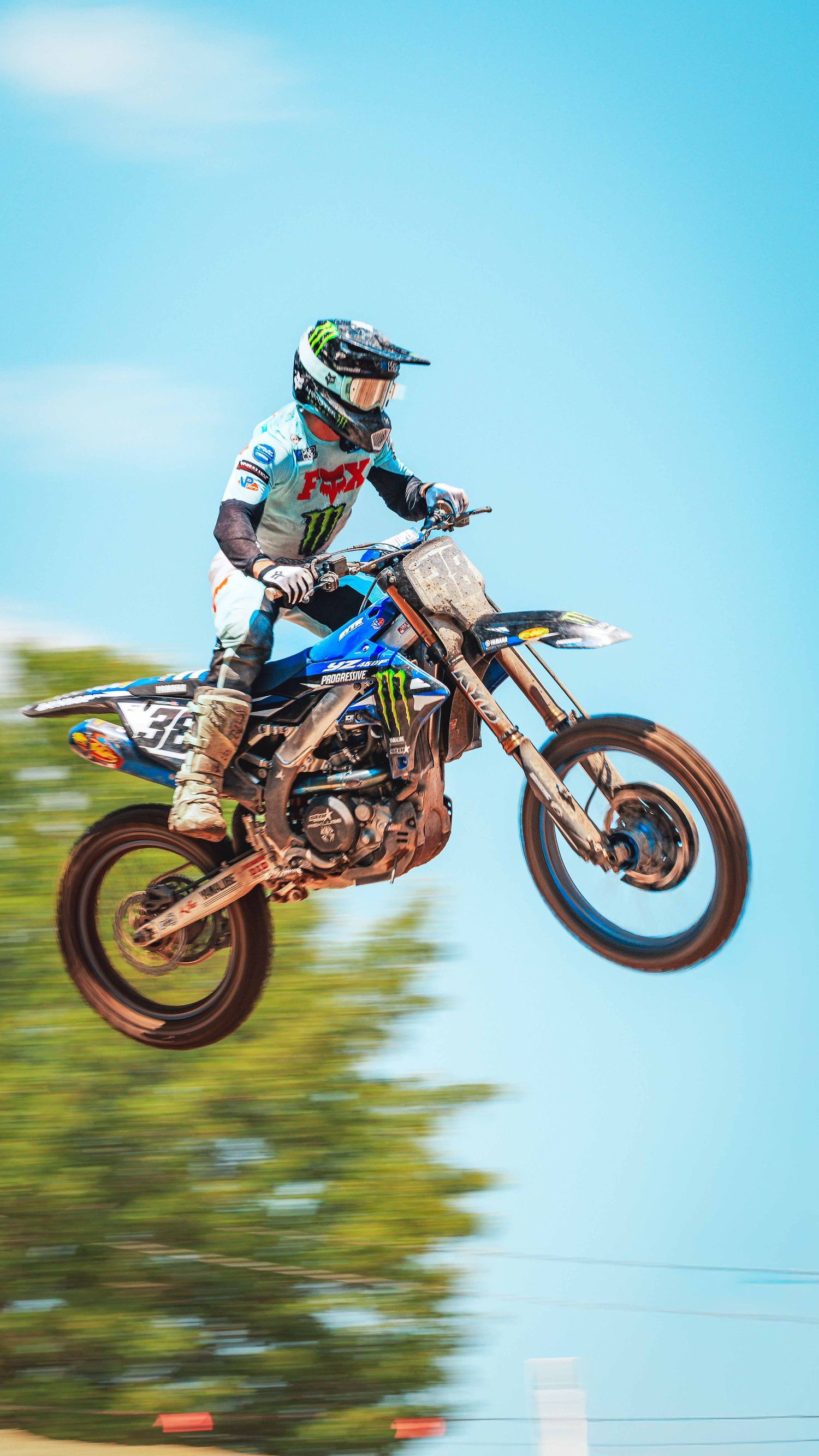 Motocross rider in Monster Energy gear performs a high jump on a blue dirt bike against a clear sky.