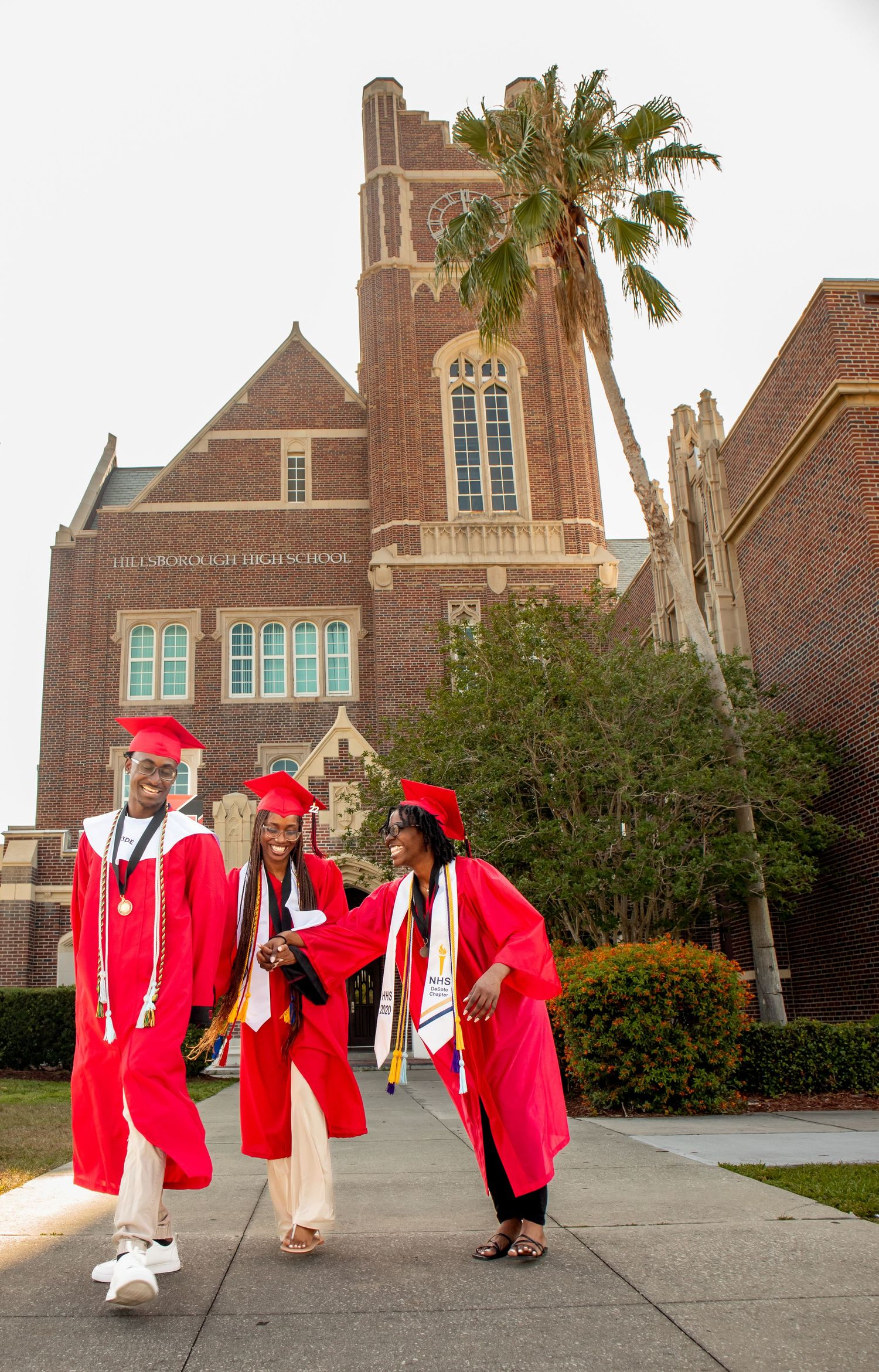 Three graduates in bright red regalia chat outside historic brick university building with palm tree.