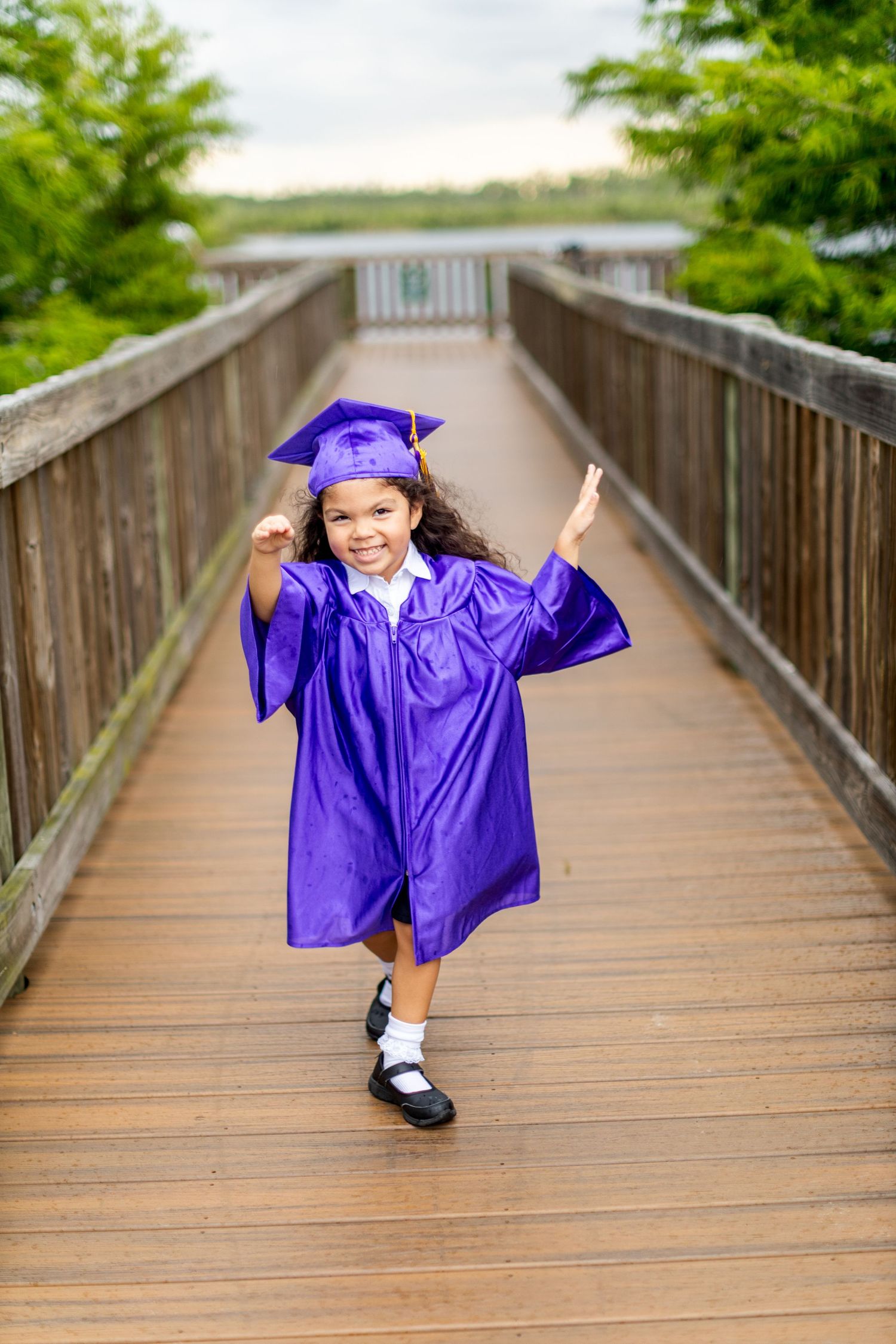 Child in purple graduation cap and gown runs playfully across wooden boardwalk.