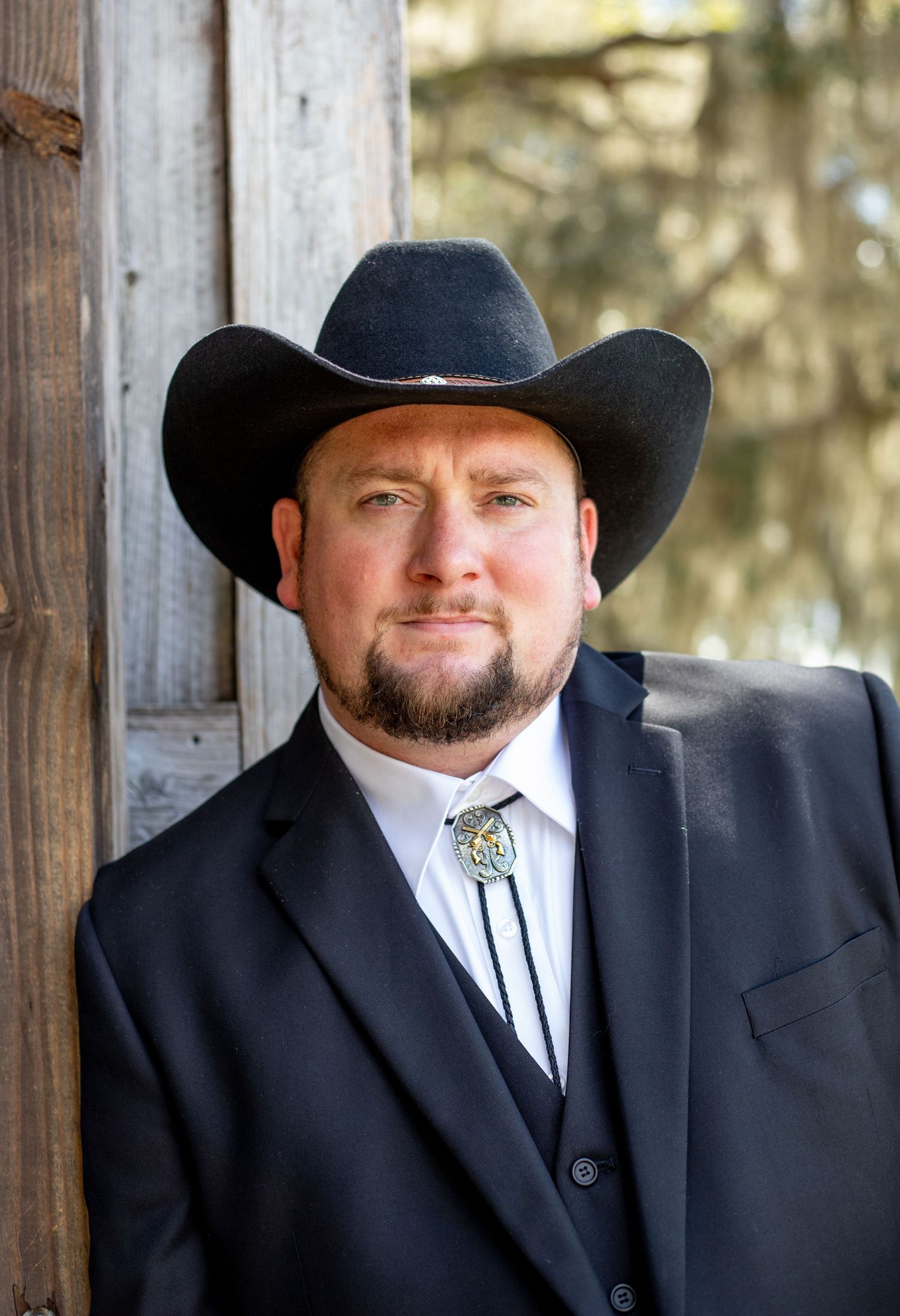 Groom in western formal wear and cowboy hat poses against rustic wooden backdrop.