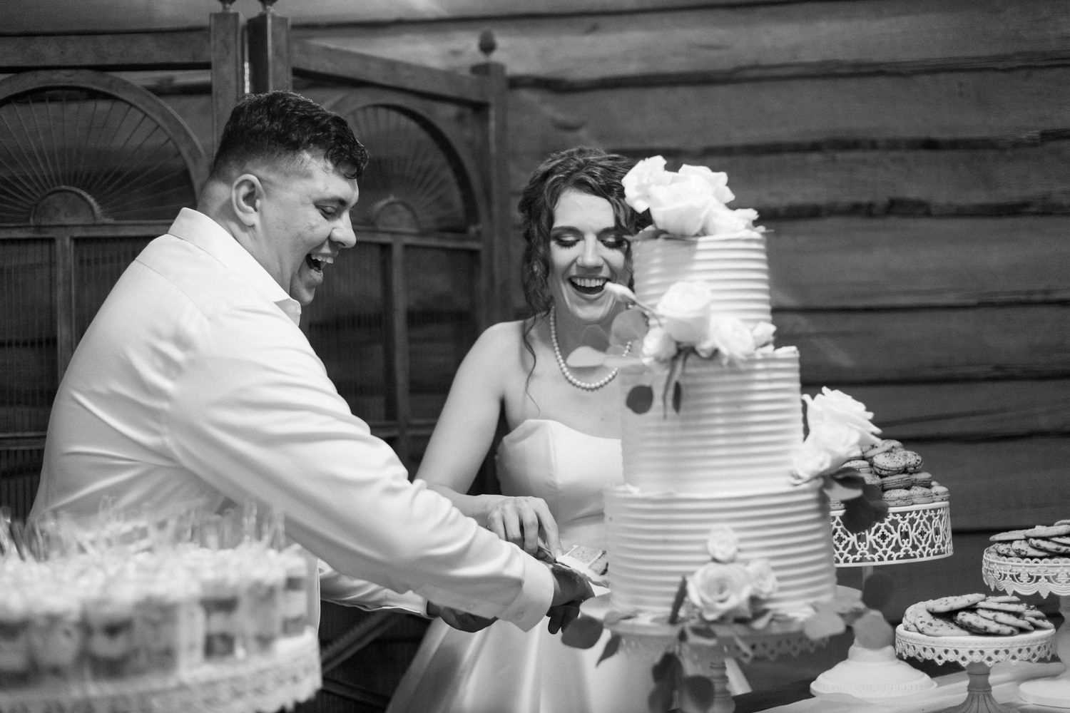 Wedding cake cutting moment at rustic wooden venue with tiered white cake.