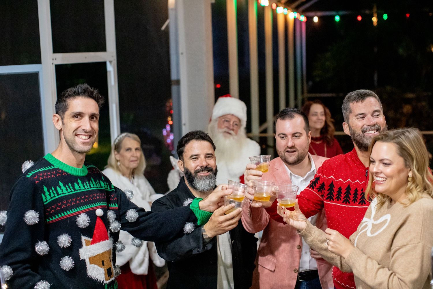 Group wearing festive sweaters raises glasses for a holiday toast with Santa nearby.