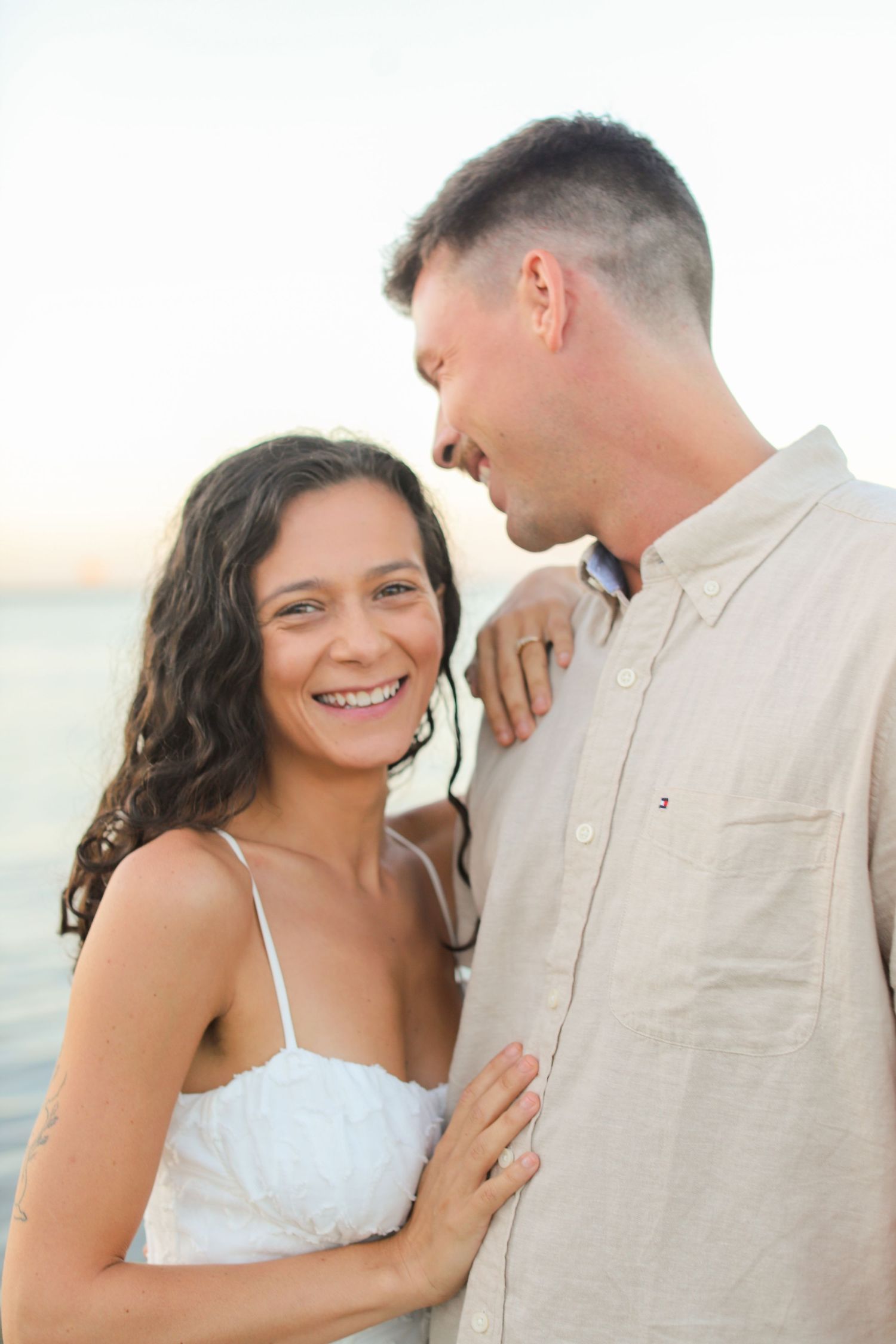 A smiling couple poses near the ocean at sunset, woman in white dress, man in beige shirt.