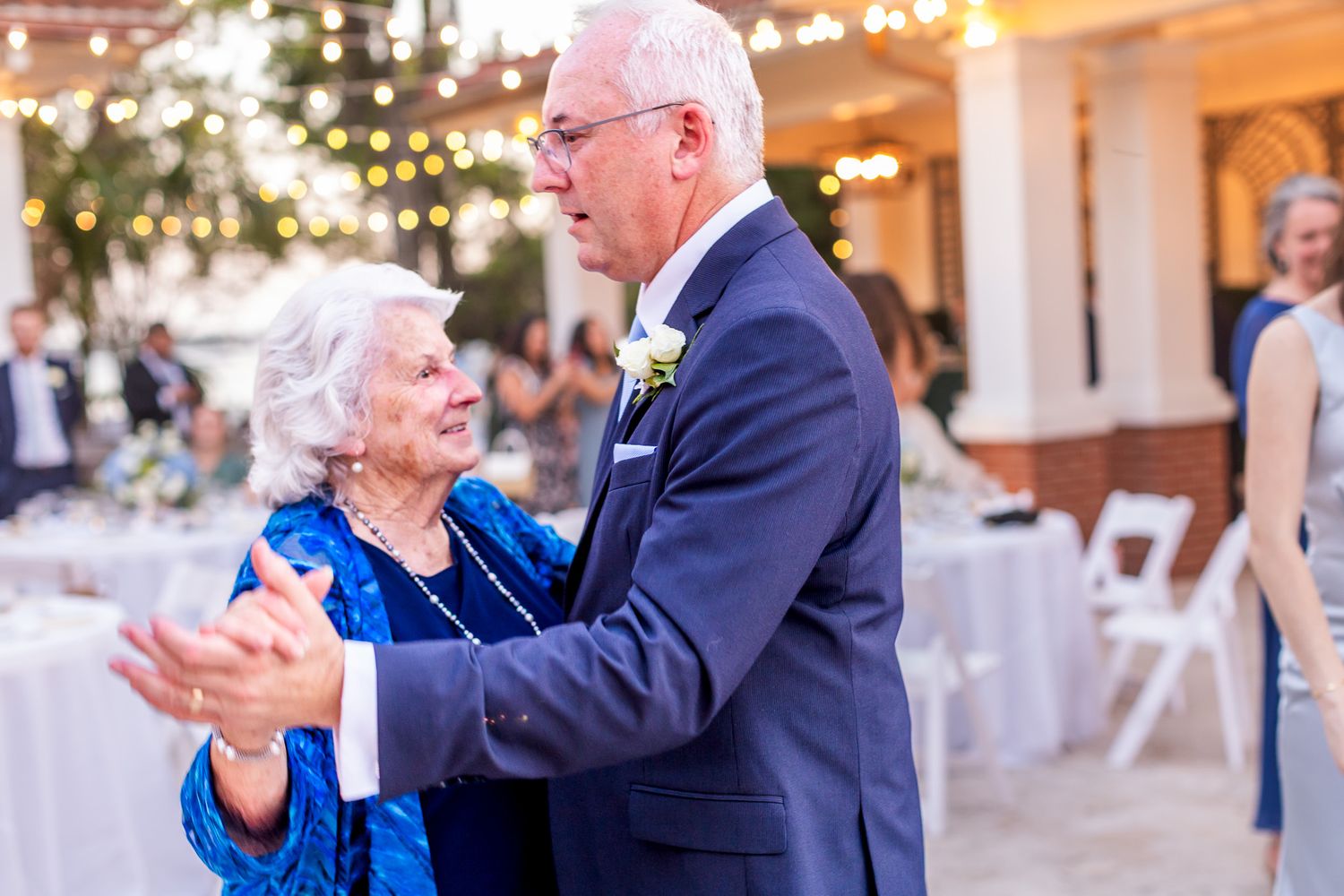 Elderly couple dancing together at an outdoor wedding reception with string lights and guests in background.