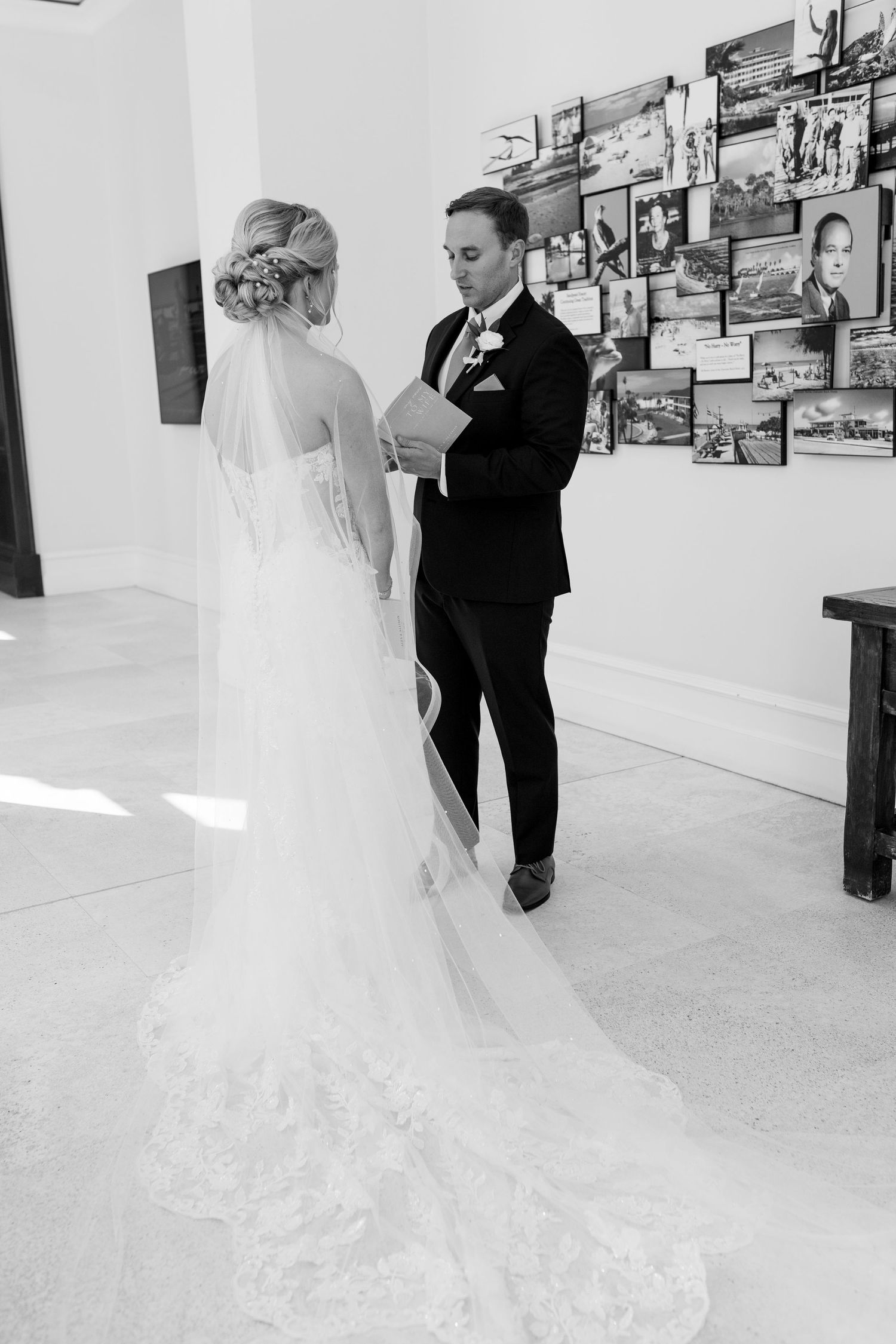 Black and white photo of a bride in a lace gown and veil facing a groom in a tuxedo during a wedding first look.