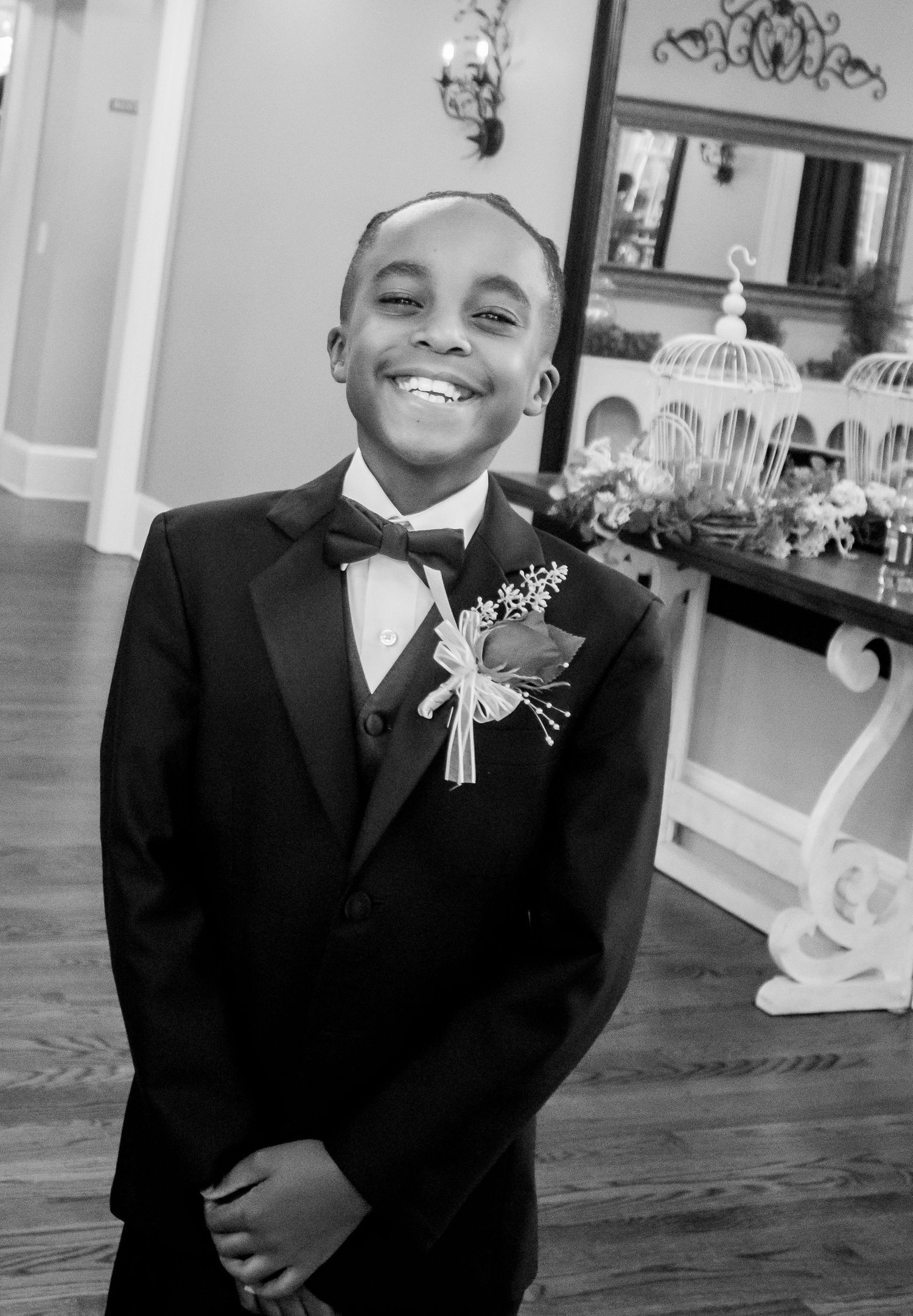 Young boy in tuxedo with boutonniere smiles at wedding ceremony in black and white photo.