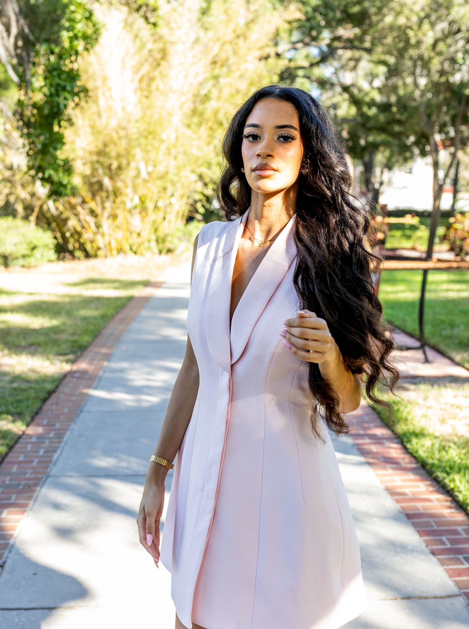 Woman in pink sleeveless blazer dress with long wavy hair poses on a sunlit brick pathway in a park.