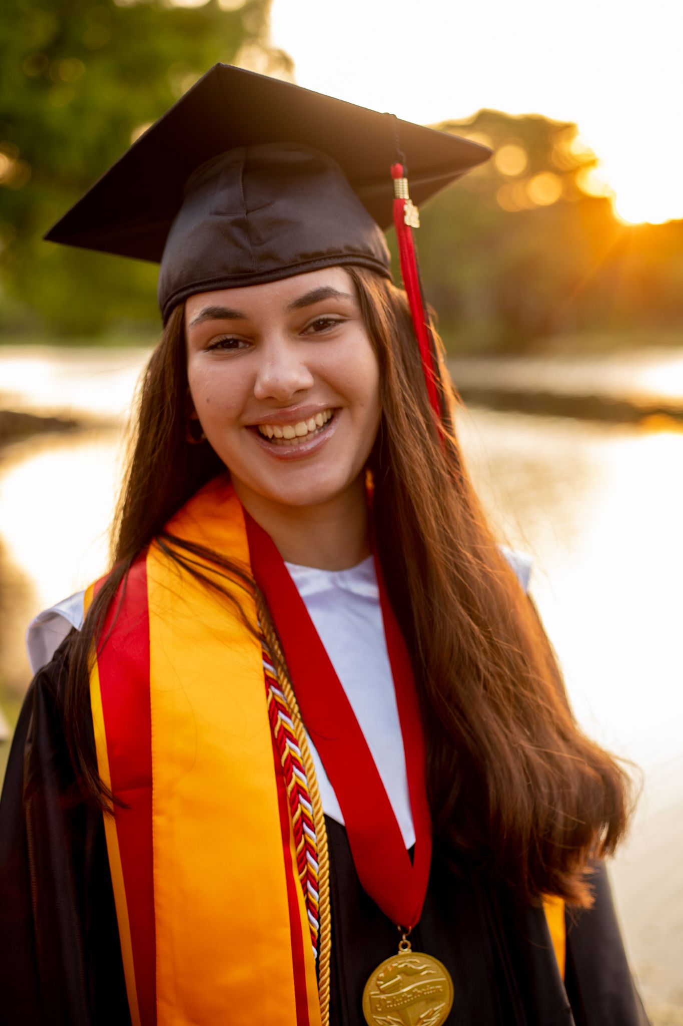 Graduation portraits showing someone in cap, gown and gold sash against sunset lighting.