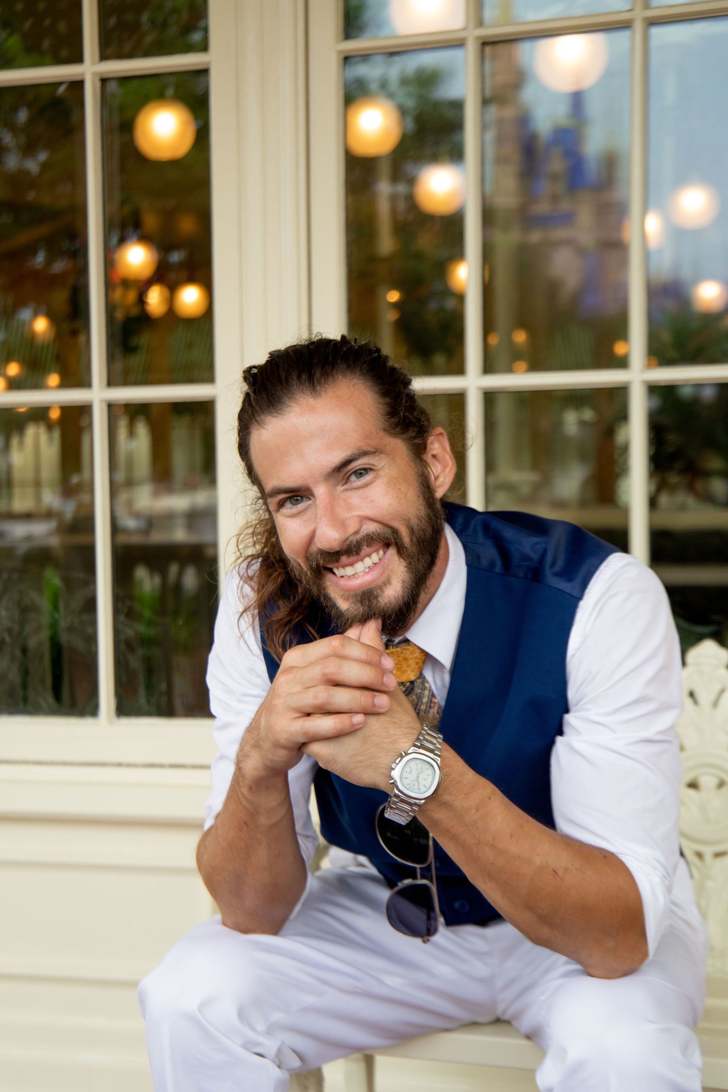 Person in navy vest and white shirt smiles while sitting by window with glowing lights.
