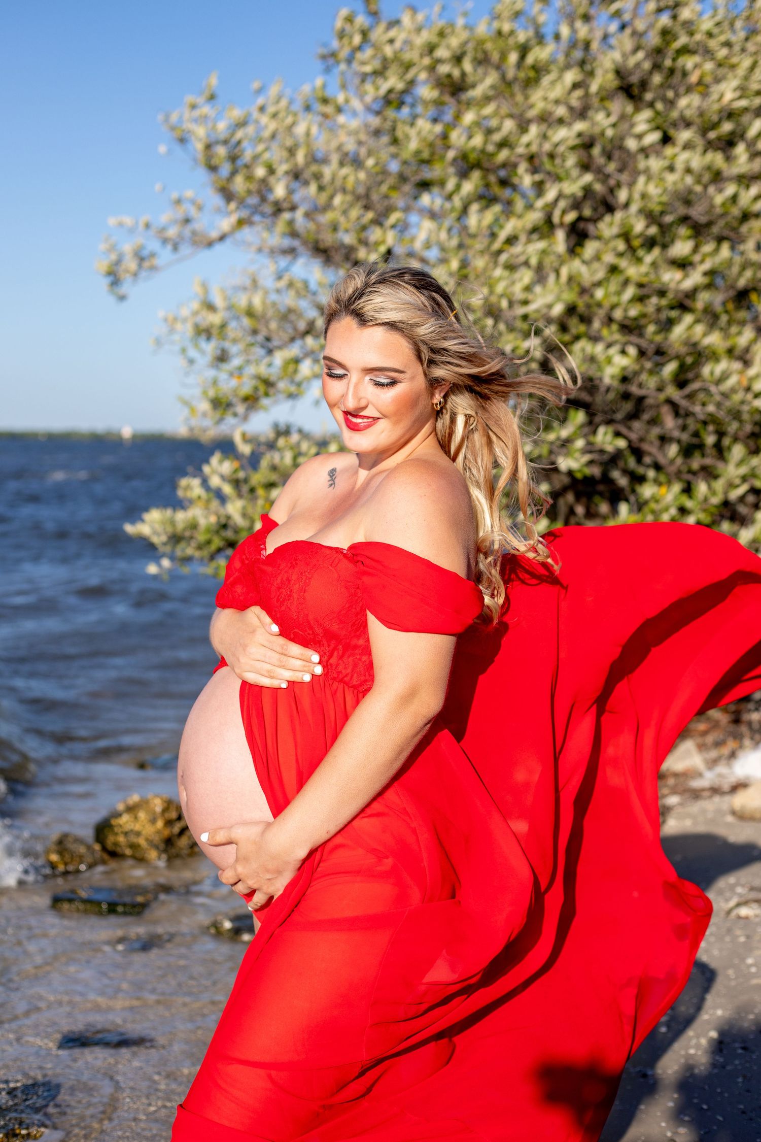 A maternity photoshoot at the beach featuring a flowing red dress against blue water and greenery.