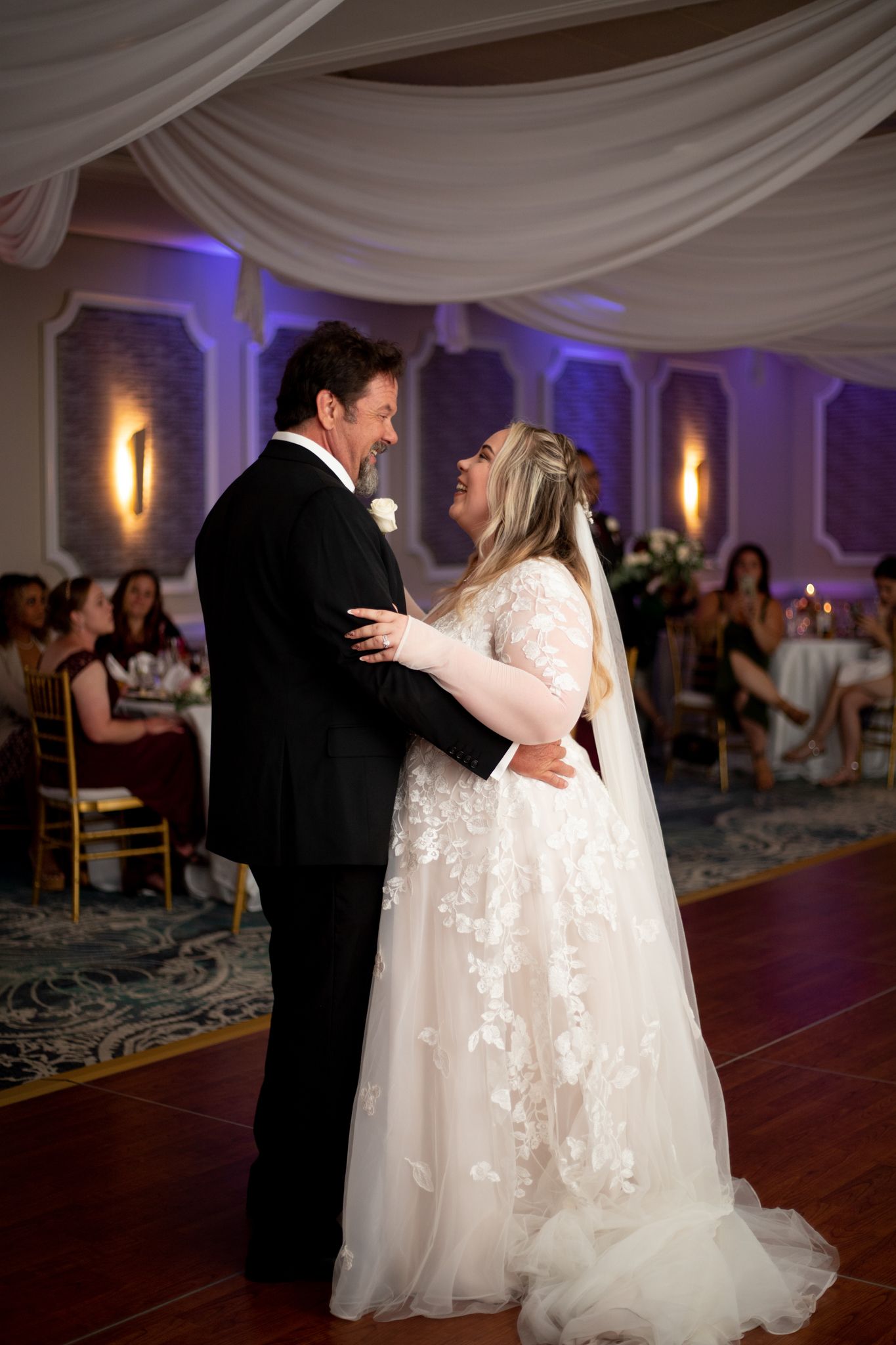 A couple shares their first dance at a wedding reception under elegant white draping and purple lighting.