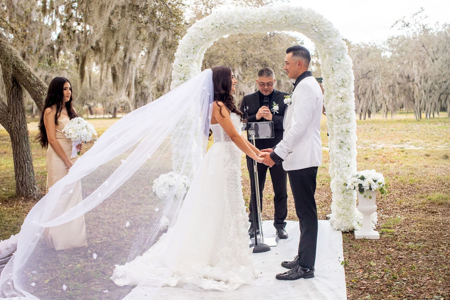 A flowing white wedding veil catches the breeze during an outdoor ceremony under oak trees.