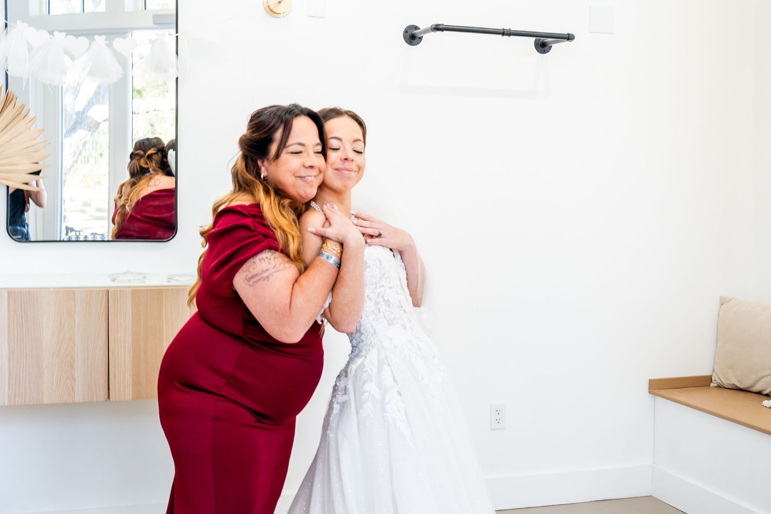 A bride in a white gown shares a tender hug with a woman in a red dress during wedding preparations.