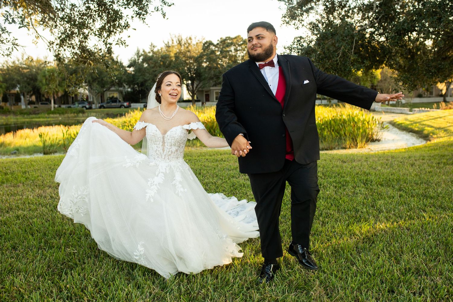 Joyful bride in white ball gown and groom in black tuxedo with red bow tie holding hands outdoors at wedding.