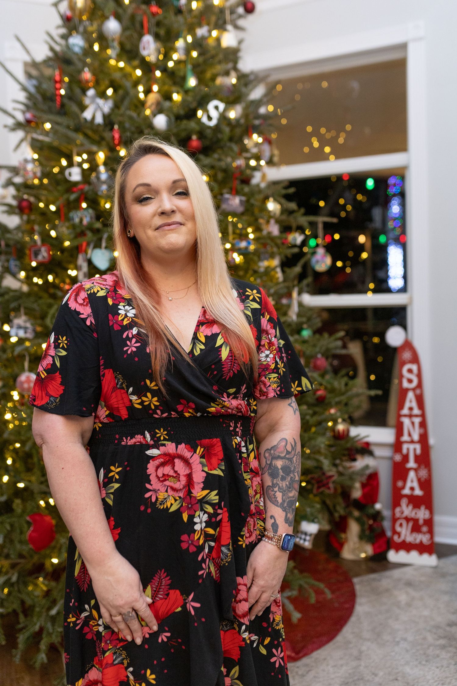 A woman in a floral dress poses in front of a decorated Christmas tree with lights.