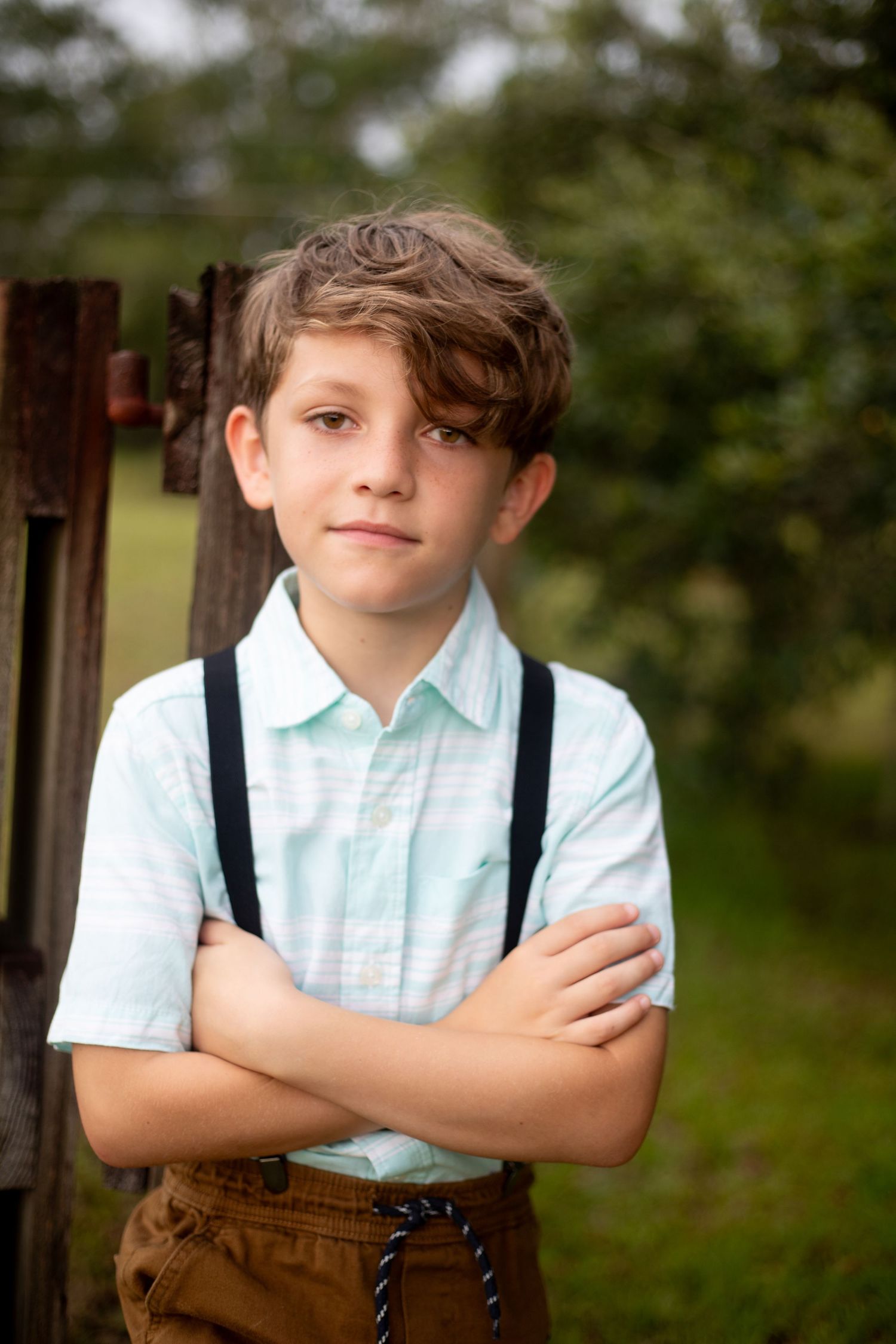 Child in light blue shirt and brown suspenders stands confidently against rustic wooden background.