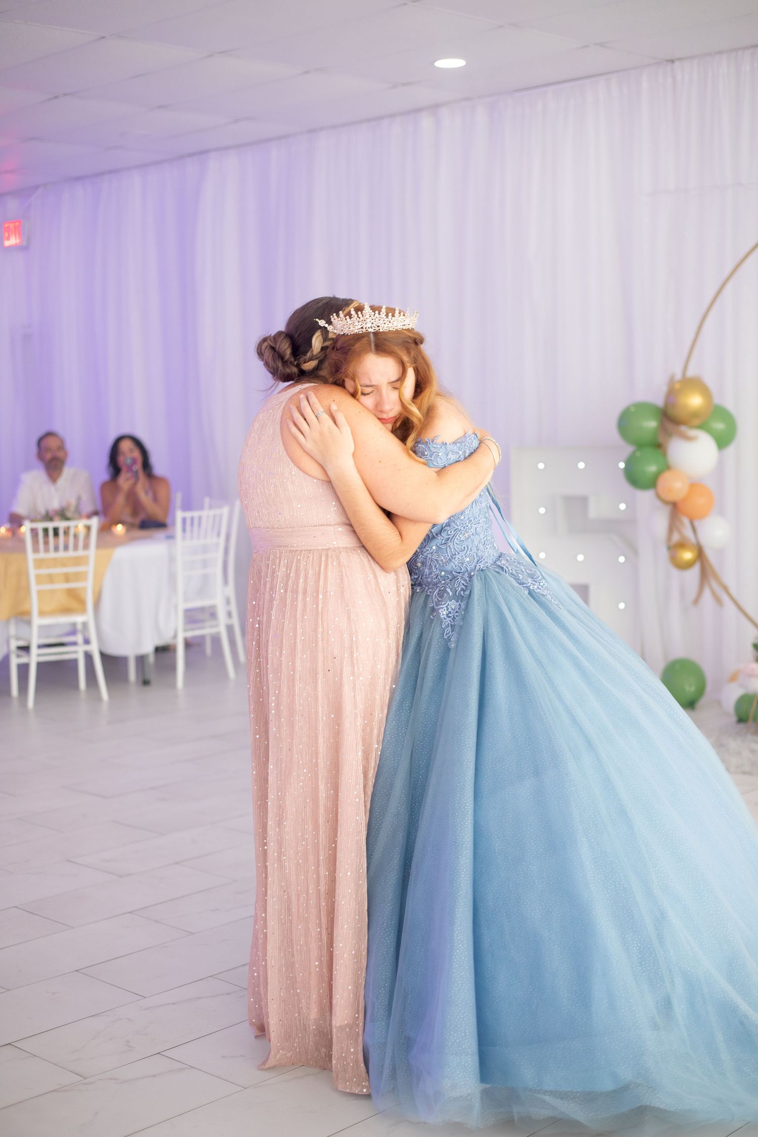 An emotional moment captured in a white-draped venue with a blue ballgown and pink dress.