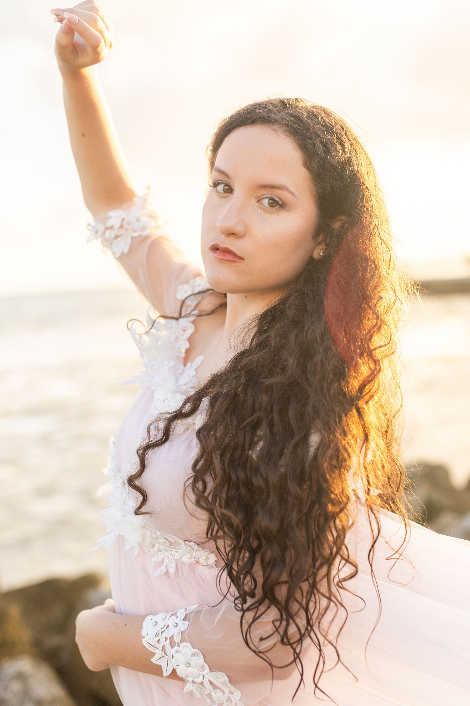Artistic beach portrait with white lace dress and natural curls in warm sunset lighting.