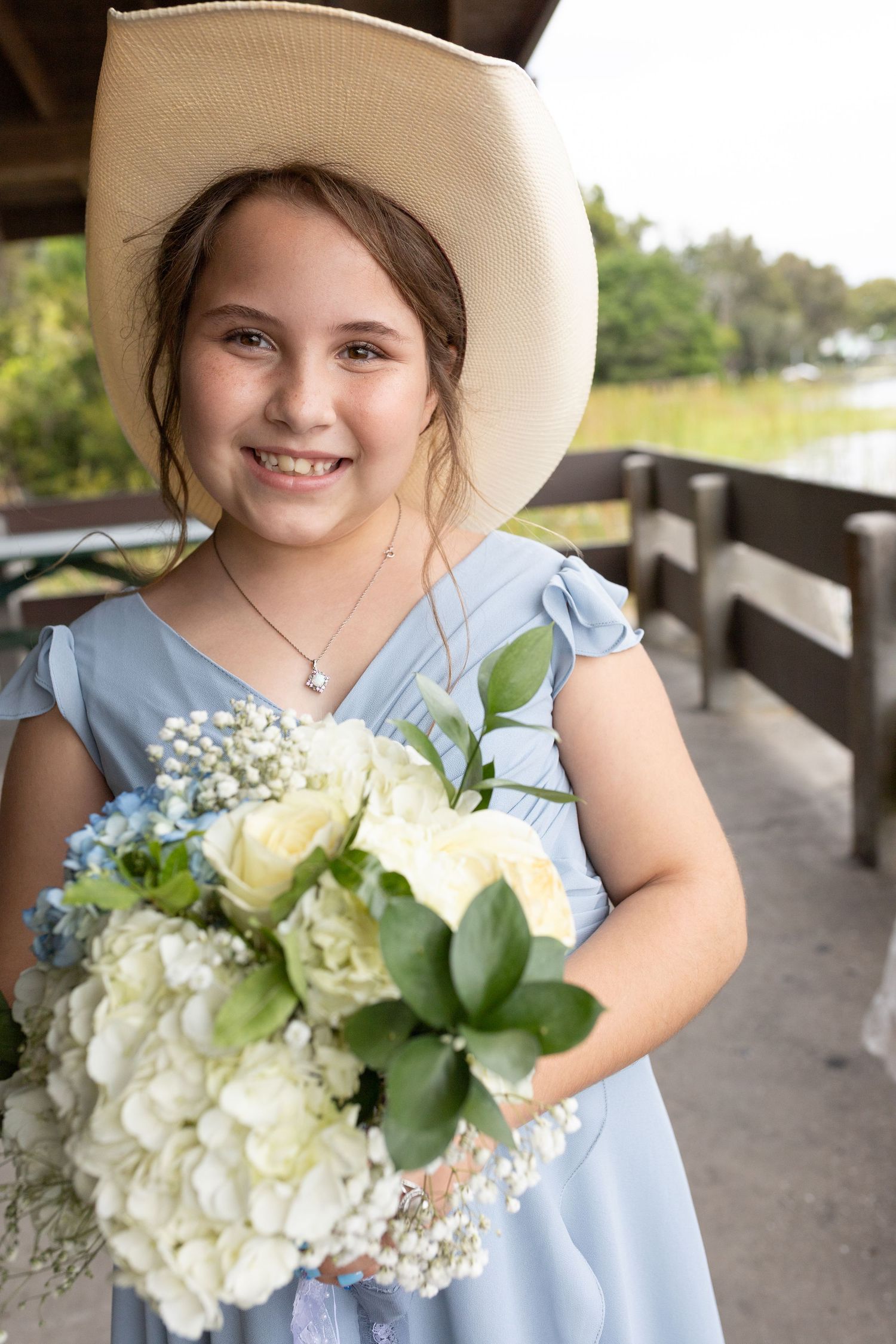 Young girl in blue dress and cowboy hat smiling while holding white hydrangea bouquet on wooden bridge.