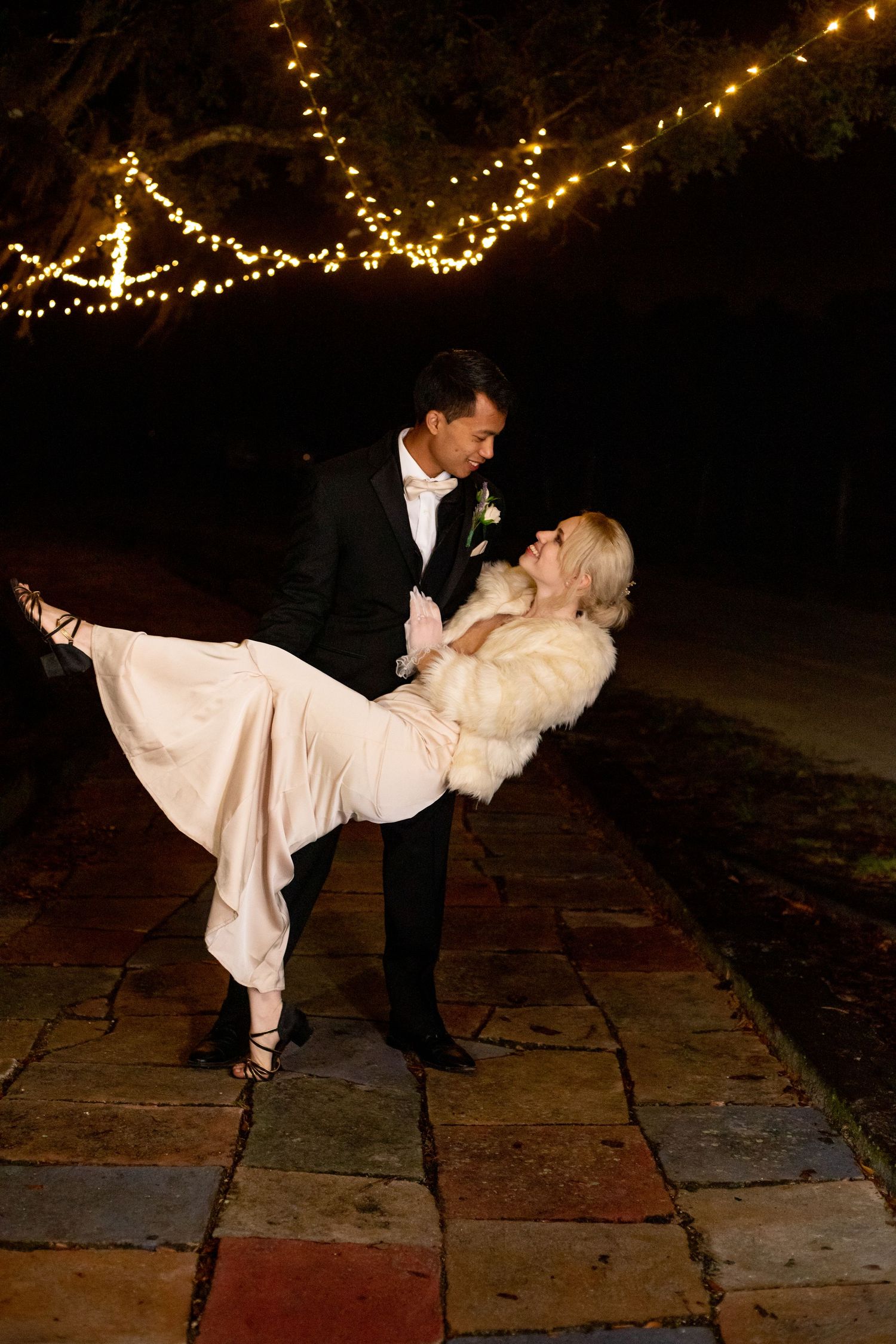 Couple sharing romantic dip dance outdoors at night under twinkling string lights, bride in blush gown and fur wrap.