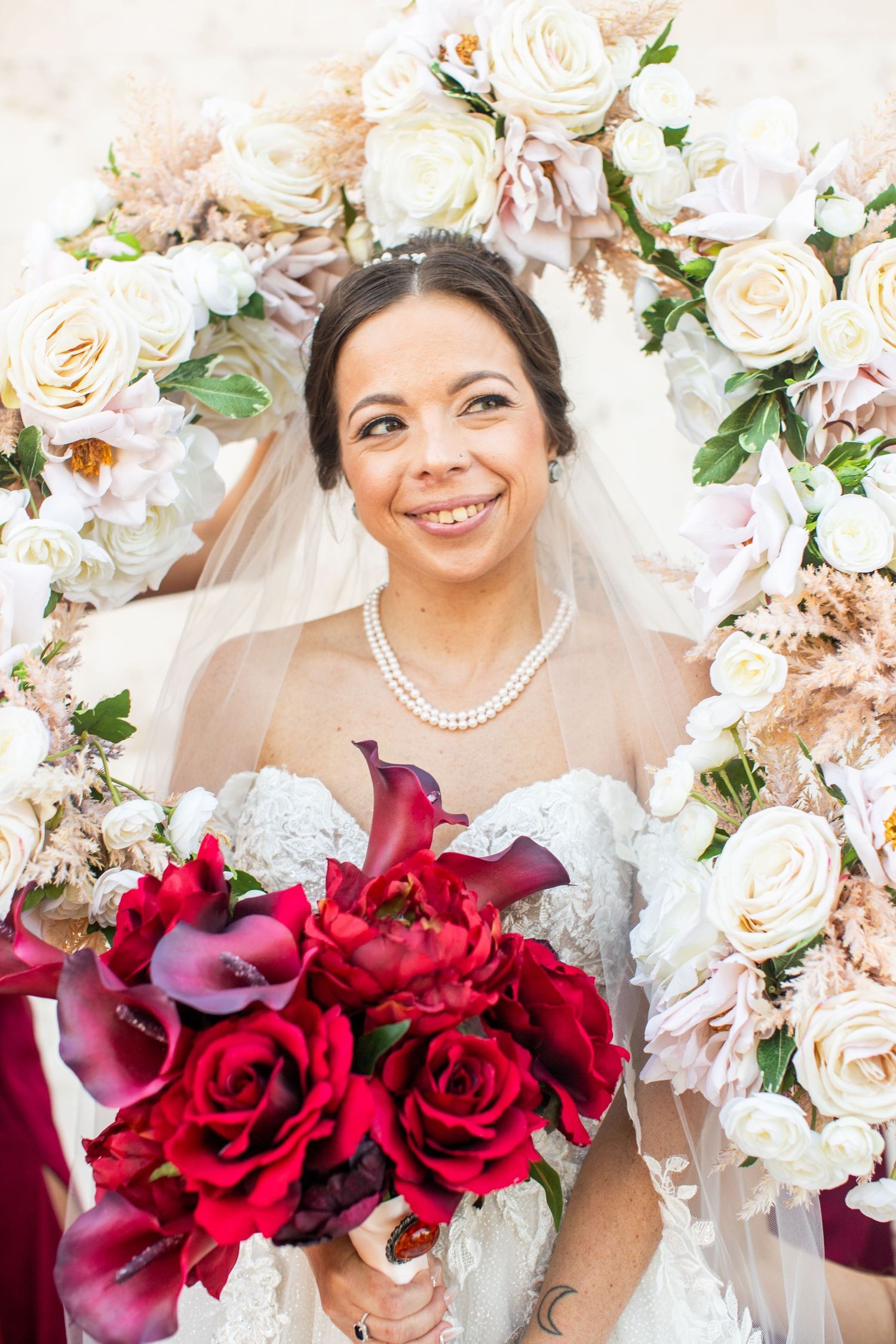 Smiling bride in lace gown holds red rose bouquet before floral arch of white and blush roses on wedding day.