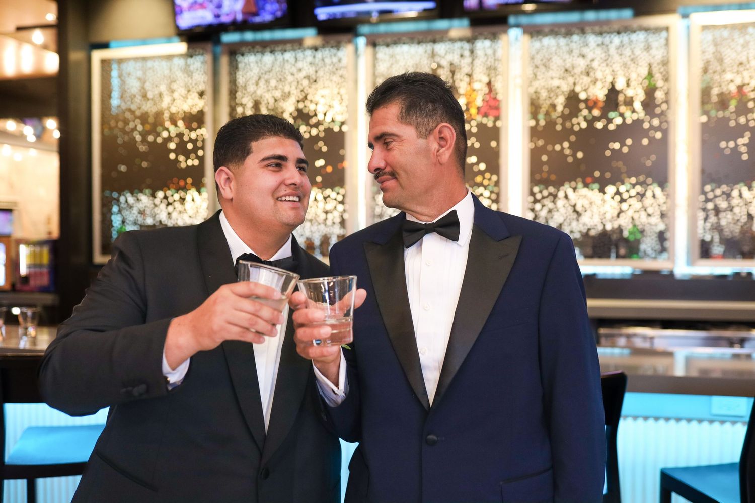 Two men in tuxedos toasting with drinks at a bar, smiling joyfully during a formal celebration.