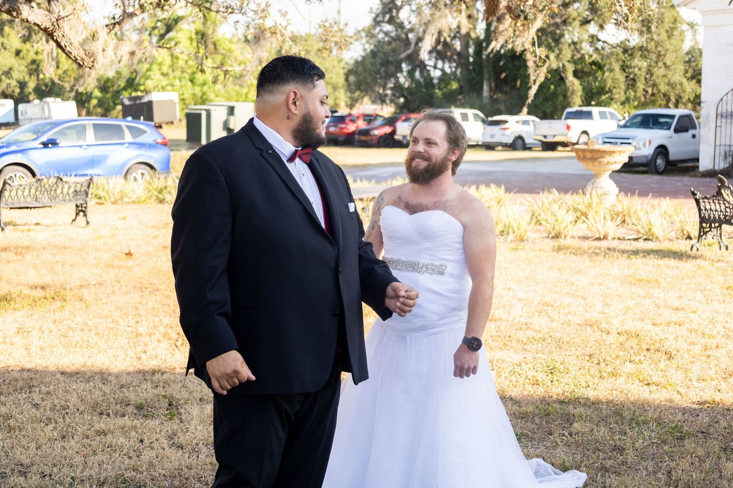 Two grooms share a tender moment outdoors, one in a tuxedo and one in a white wedding gown, on their wedding day.