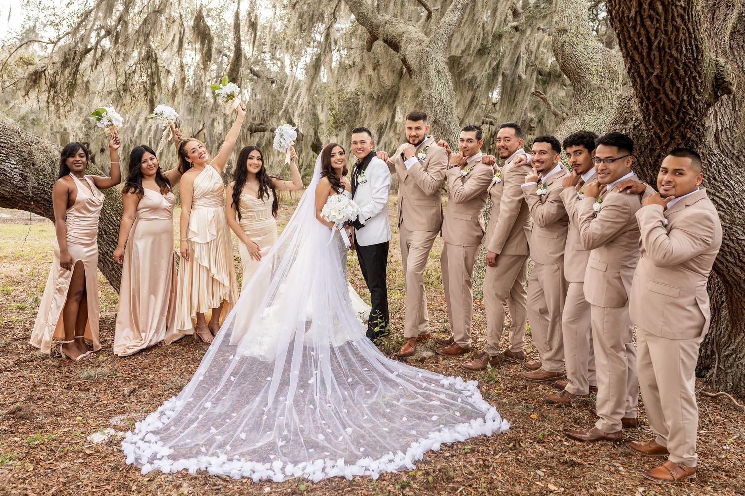 Wedding party members in beige and white attire gather under Spanish moss trees.