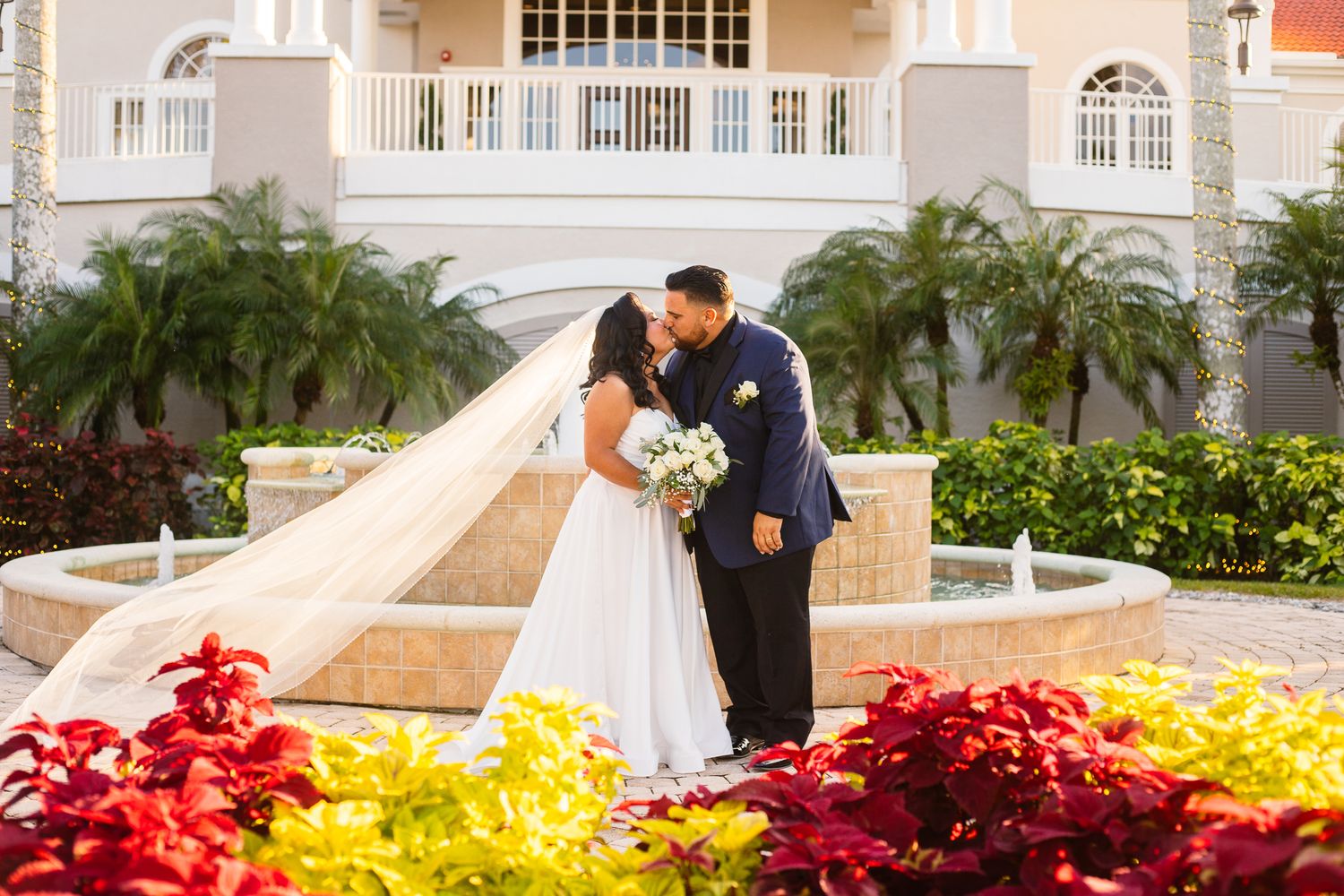 Wedding couple embraces near colorful floral arrangements and white architecture at sunset.