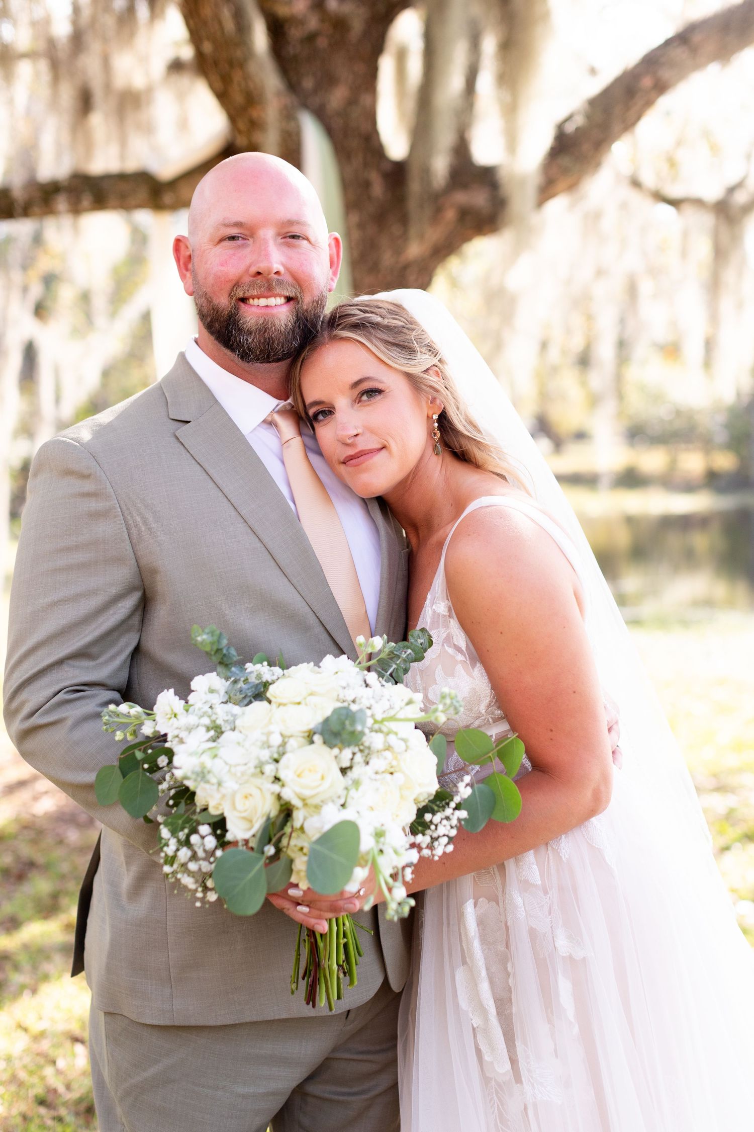 Wedding couple poses under moss-draped trees with white floral bouquet and gray suit.
