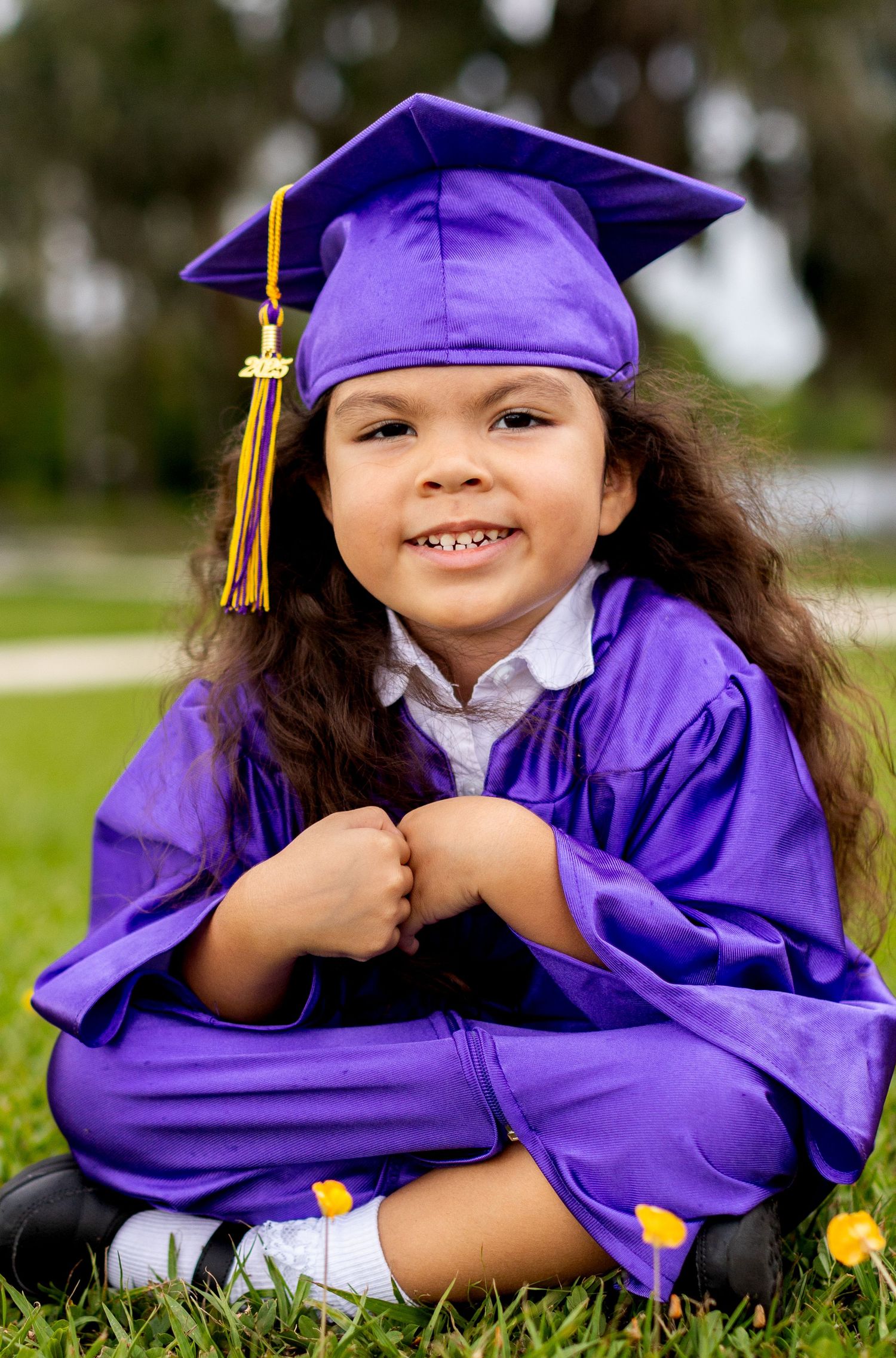 Graduate in purple cap and gown with gold tassel sits smiling on grass.