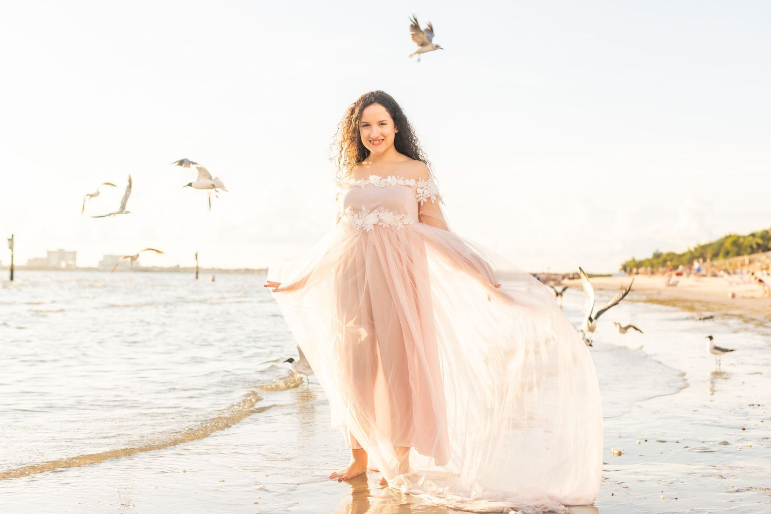 Romantic beachside portrait in a flowing blush gown with seabirds in flight.