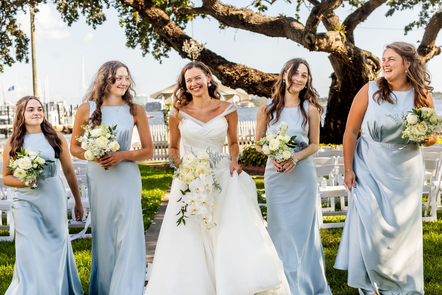 Bride in white gown walks with four bridesmaids in light blue satin dresses holding white bouquets outdoors.