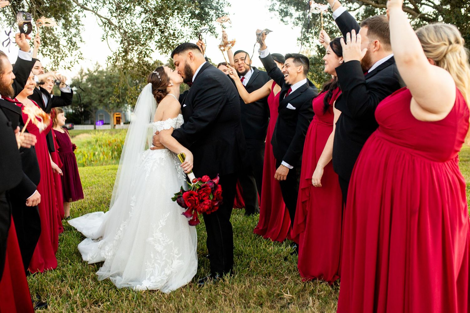 Groom lifts bride during joyful wedding party celebration, bridesmaids in red gowns and groomsmen in black tuxedos cheering outdoors.