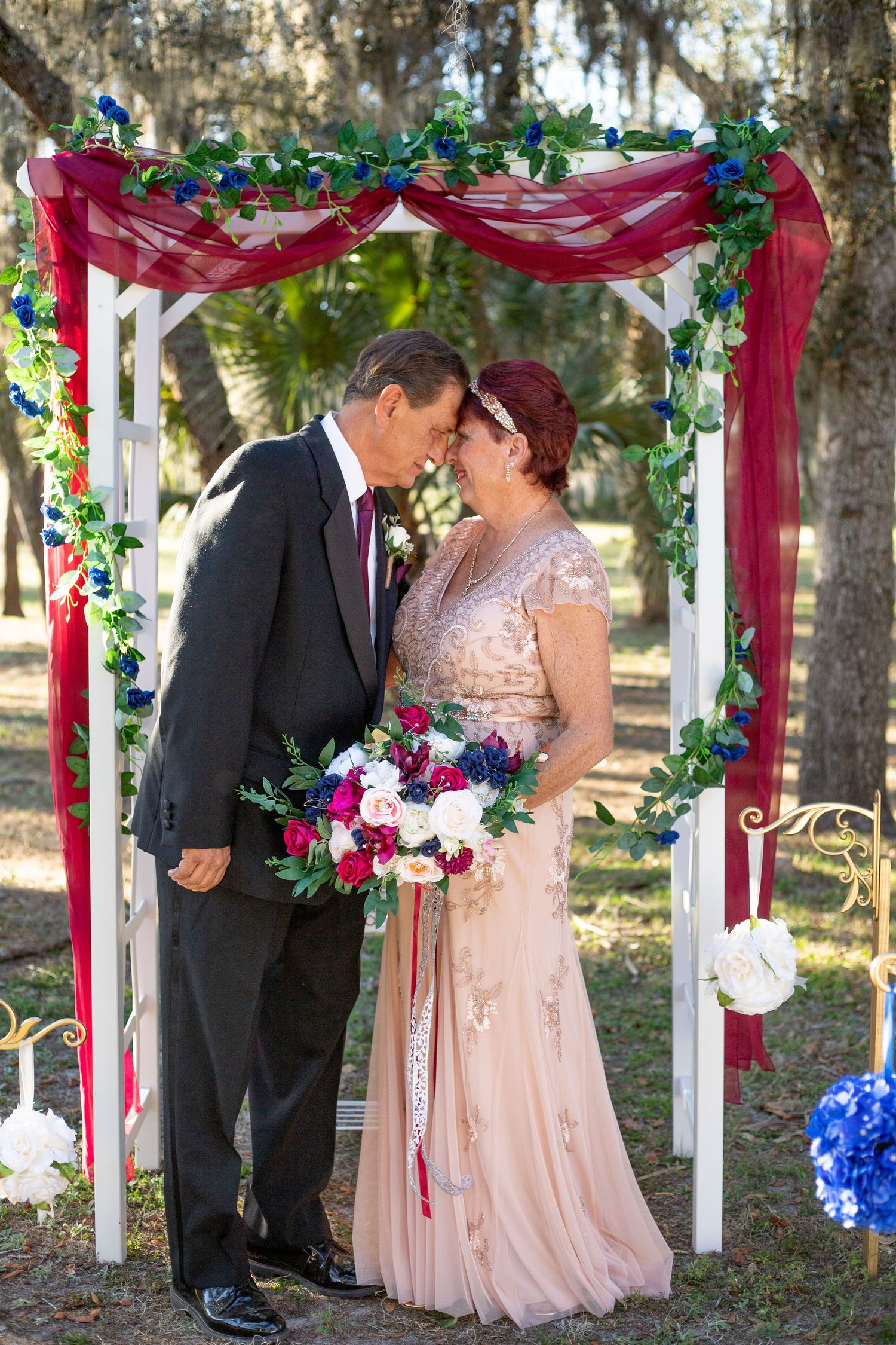 Wedding ceremony under a decorated arbor with red draping and colorful floral arrangements.