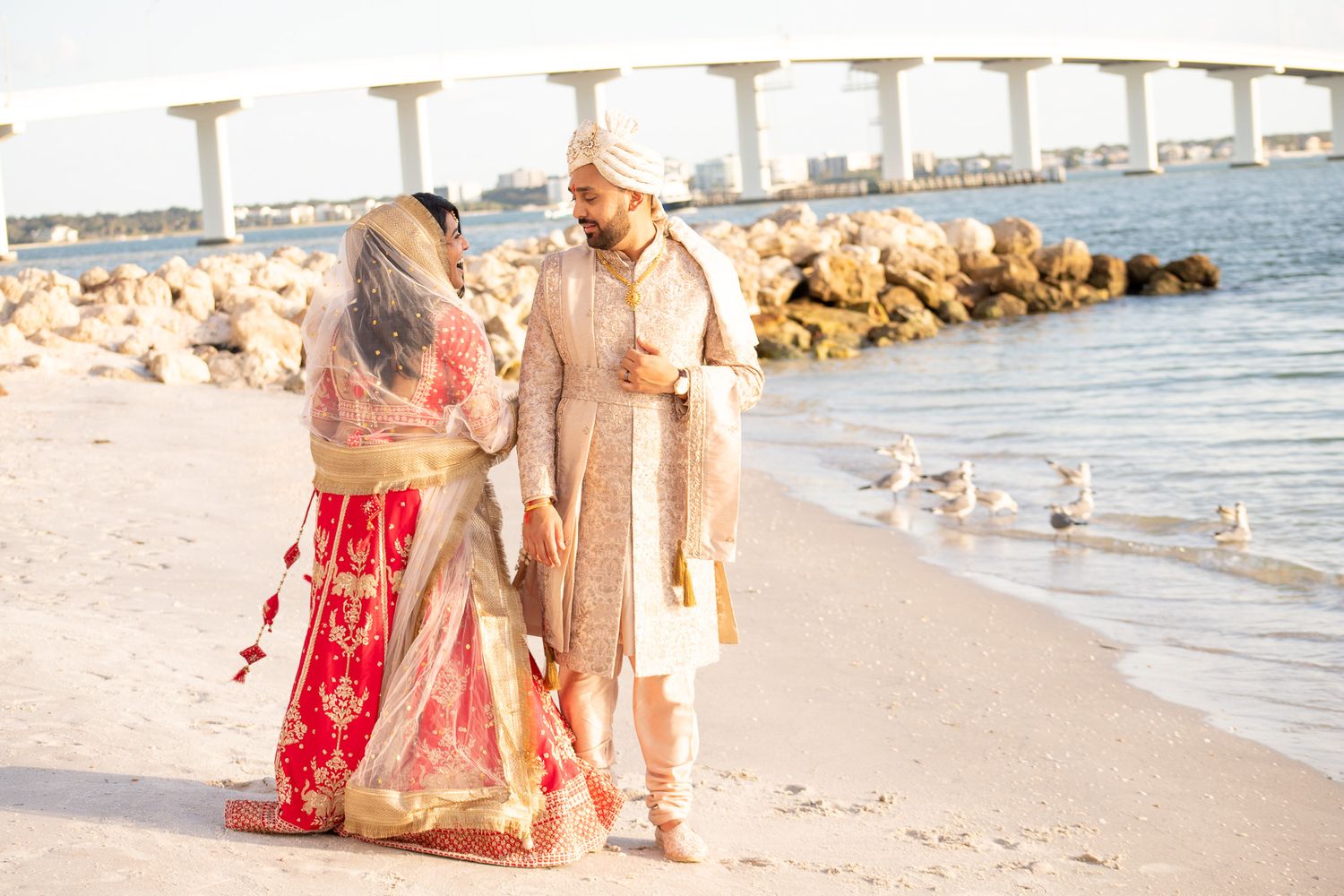 Couple in traditional Indian wedding attire walking along a beach with a bridge in the background.