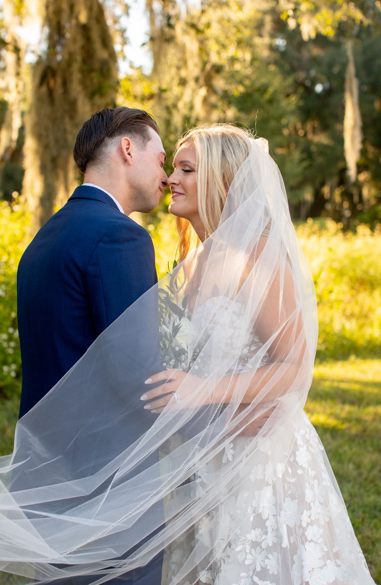 A bride and groom share a romantic almost-kiss outdoors, her lace gown and flowing veil catching golden light among lush green trees.
