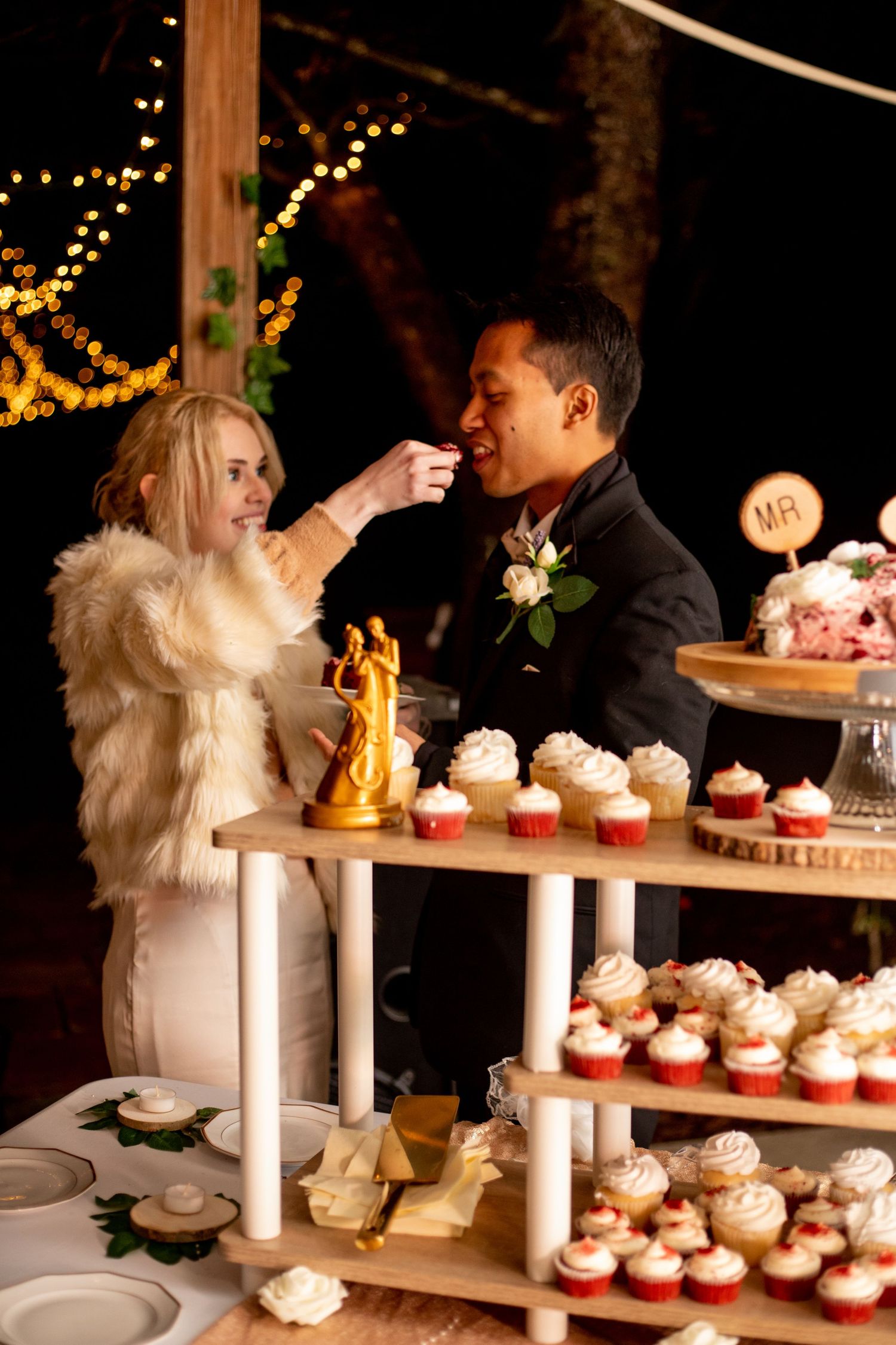 Bride and groom sharing cake at wedding reception with cupcake display tower and warm string lights.